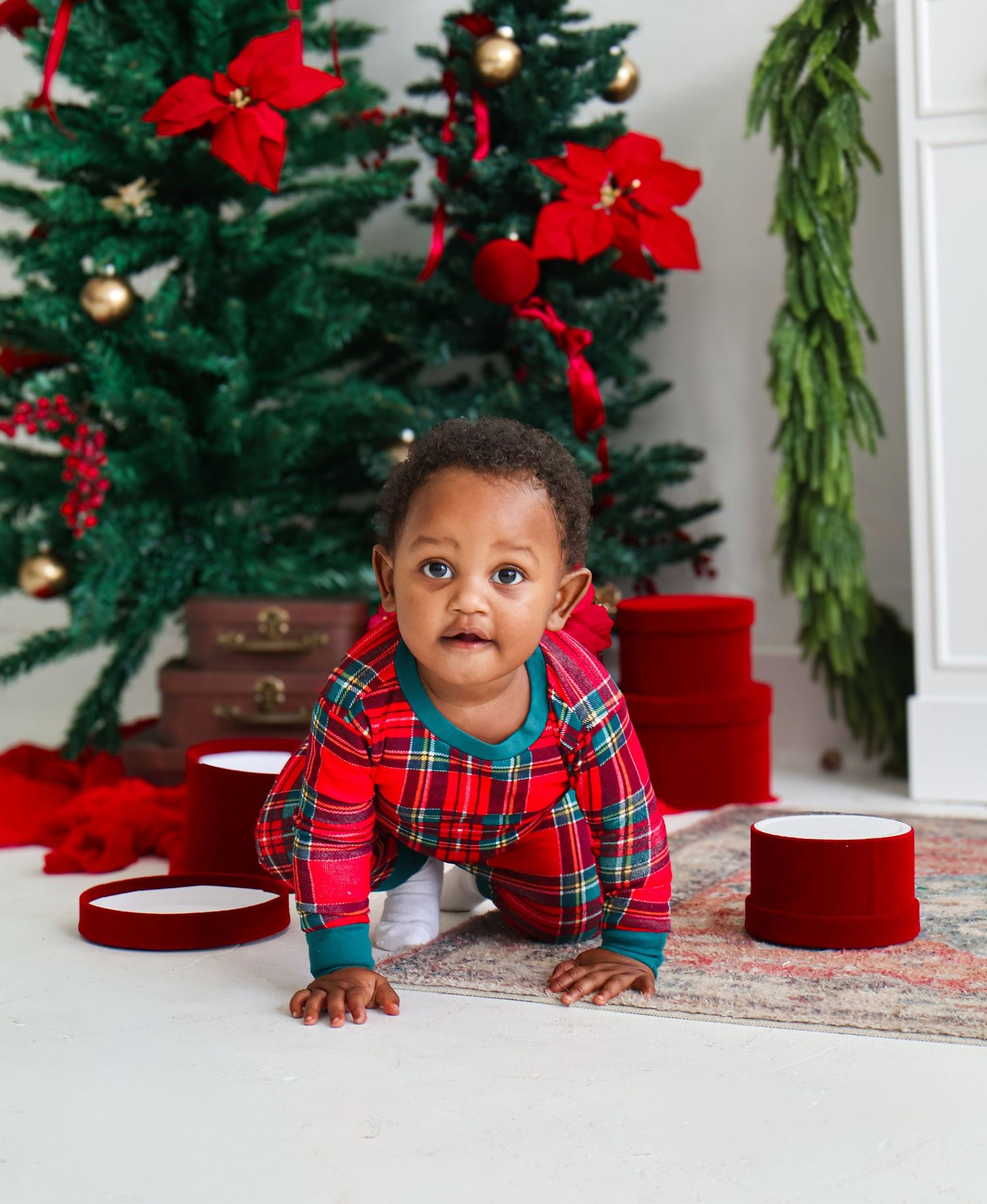 Baby in red plaid Christmas pajamas crawling on floor near decorated tree and red gift boxes.