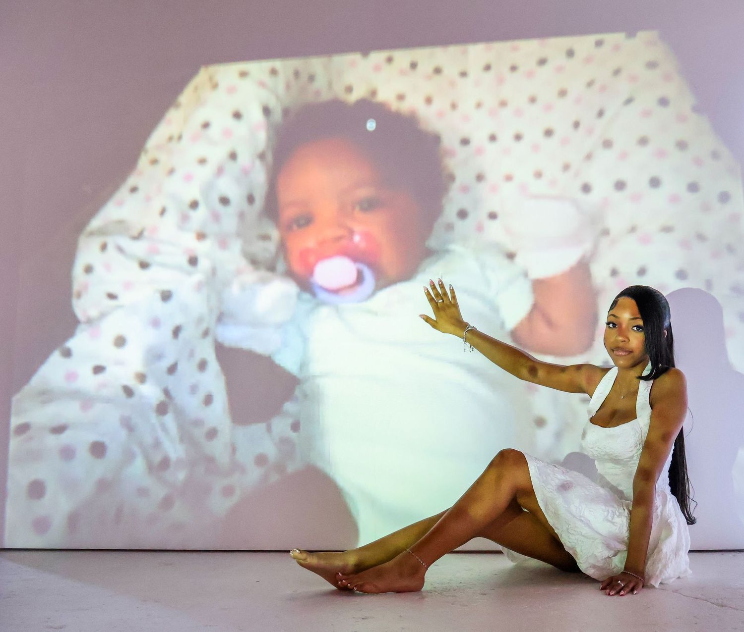 Woman in white dress poses beside large projected image of a baby with a pacifier on a gallery wall.