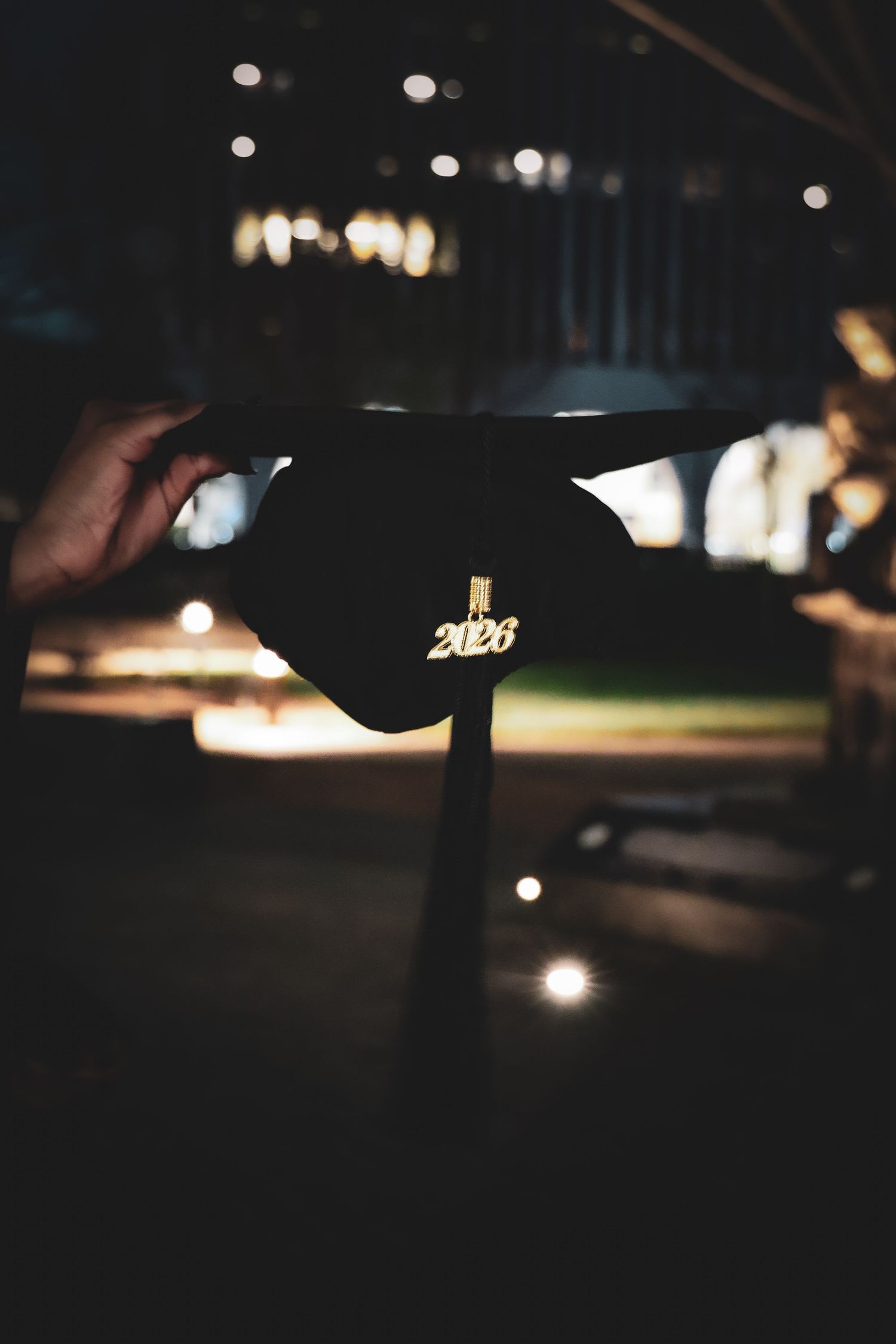 Hand holding a black graduation cap with gold 2023 charm against a dark bokeh background.