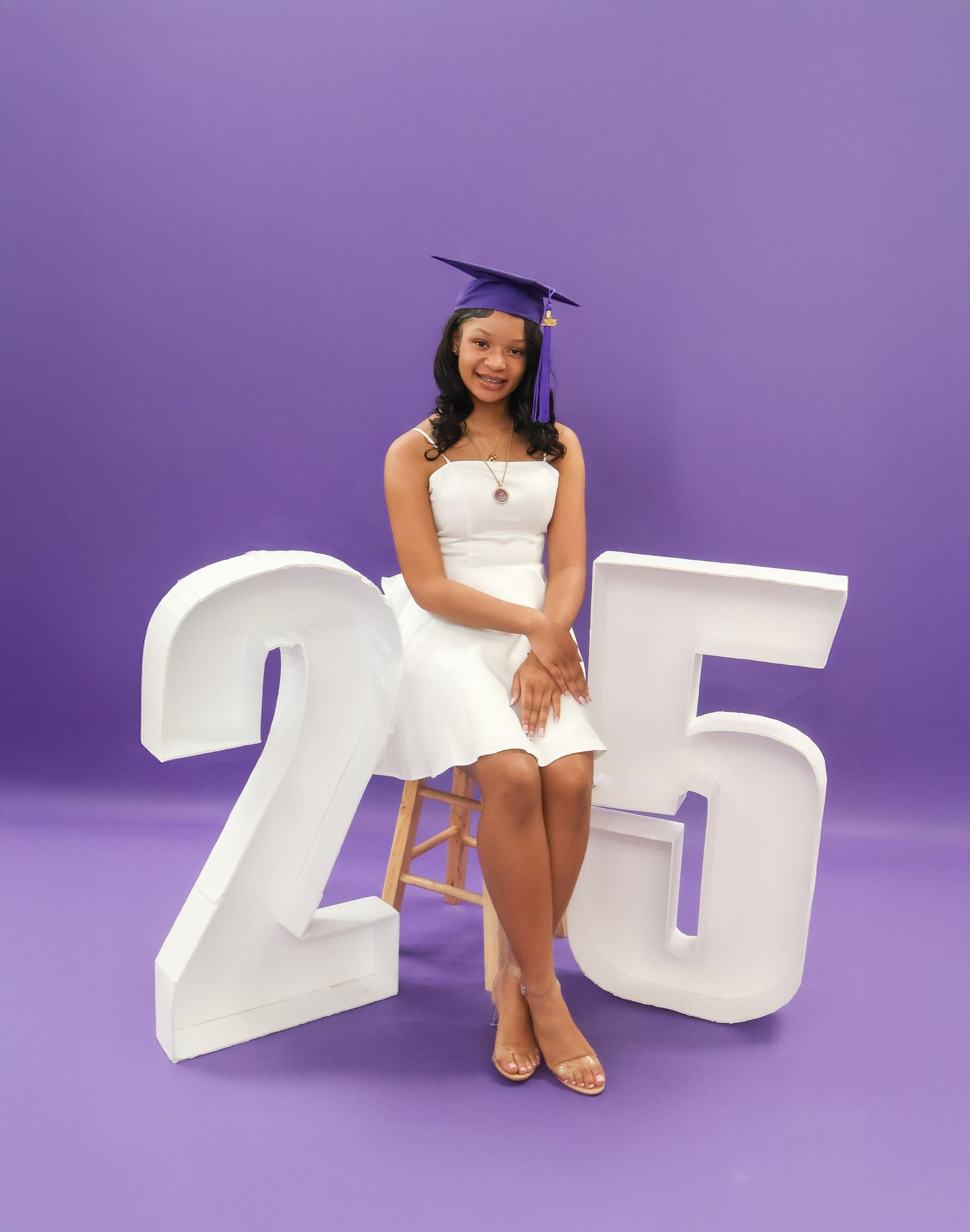 Smiling graduate in white dress poses with large '25' props on purple background.