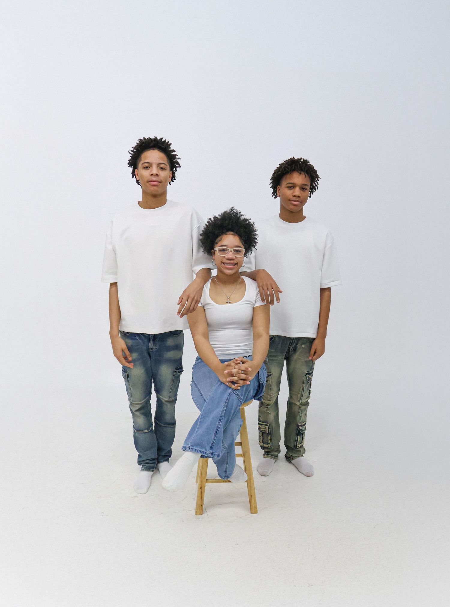 A woman sits on a stool flanked by two teenage boys, all wearing white tops and jeans against a white background.
