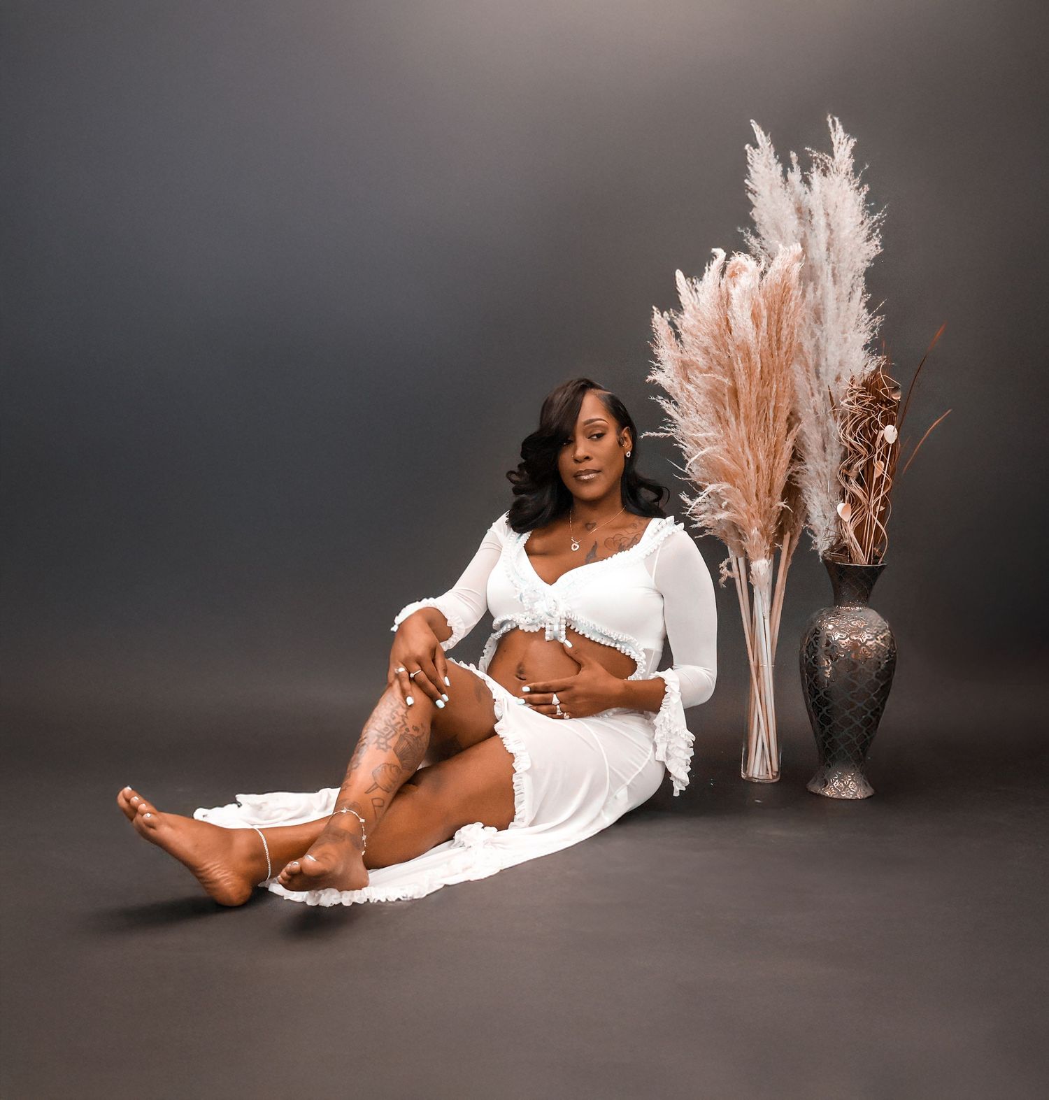 Pregnant woman in white crop top and skirt sits gracefully beside decorative vases with pampas grass.