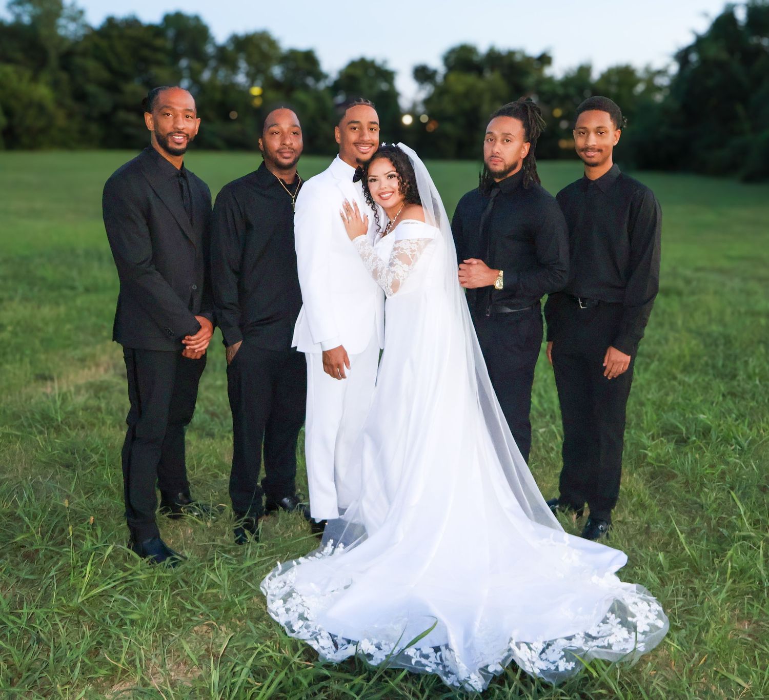 Bride in white gown and veil poses with groom in white suit and groomsmen in black attire on green lawn.