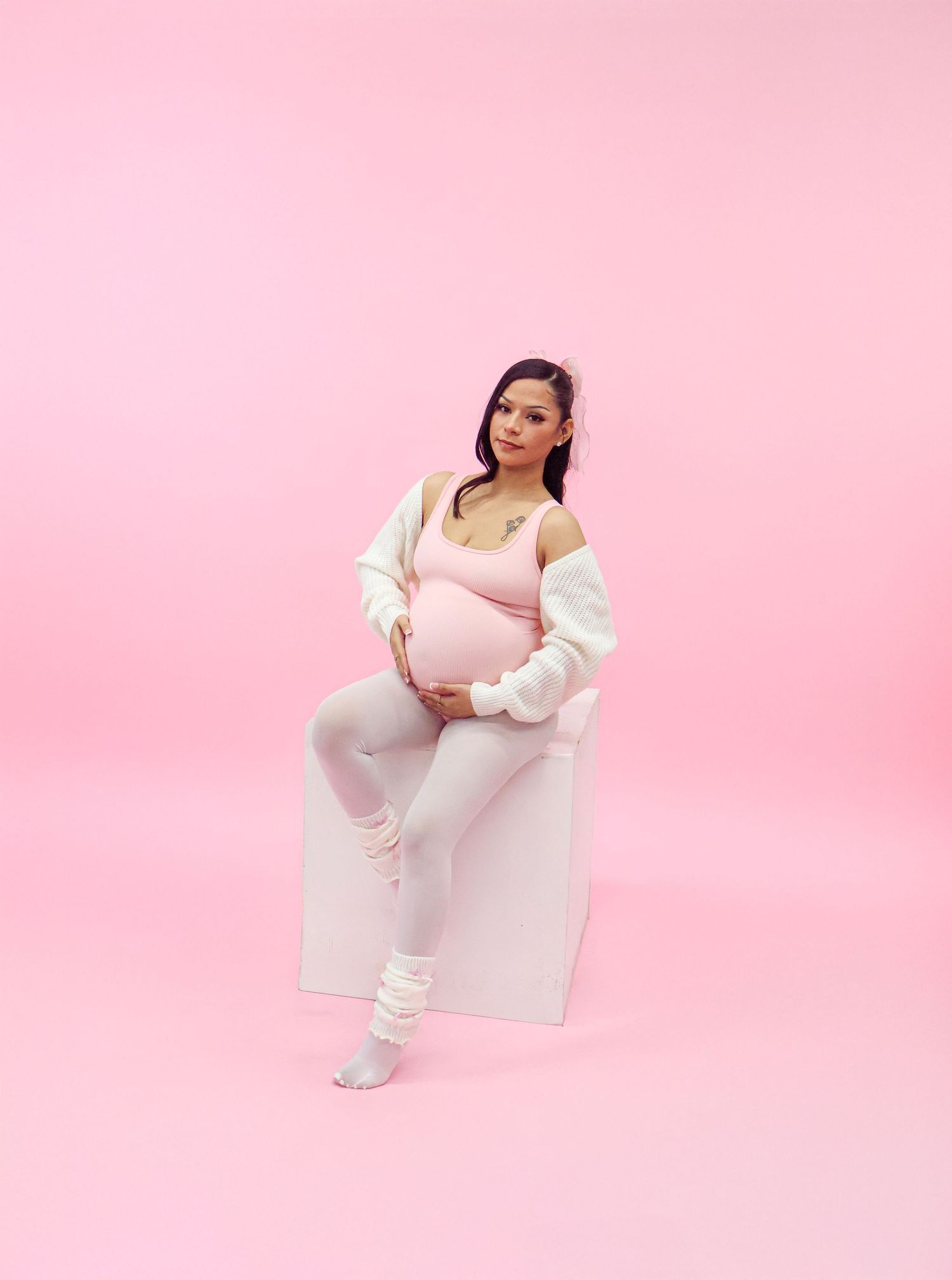 Pregnant woman in pink activewear sits on white cube against bright pink studio background.