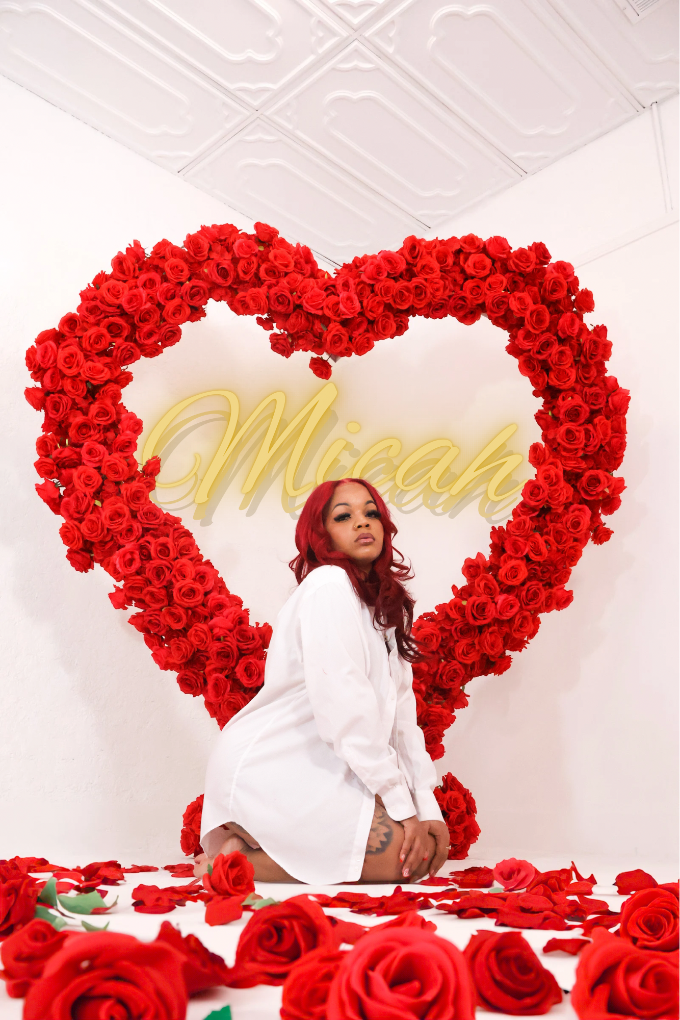 A woman in a white dress kneels before a large red rose heart wreath with a Micah neon sign.