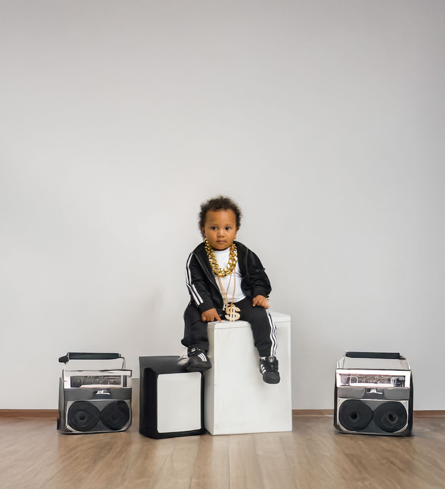Toddler in Adidas tracksuit with gold chain sits on white cube flanked by boomboxes.