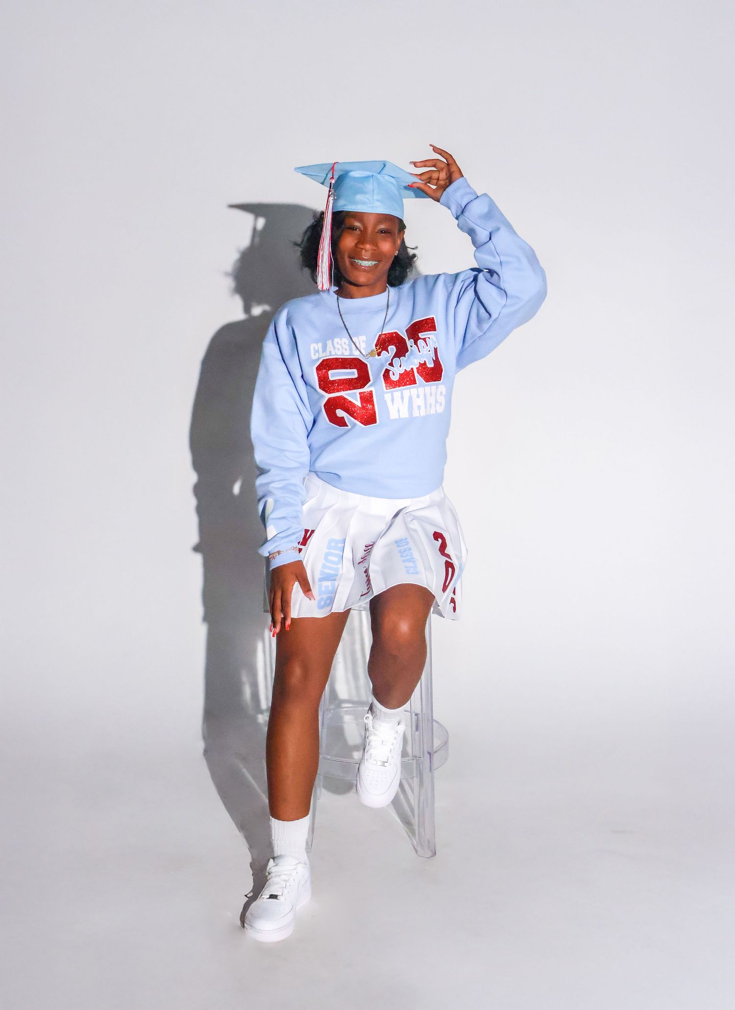 Smiling graduate in blue 2025 sweatshirt and cap poses dynamically in white shorts and sneakers.