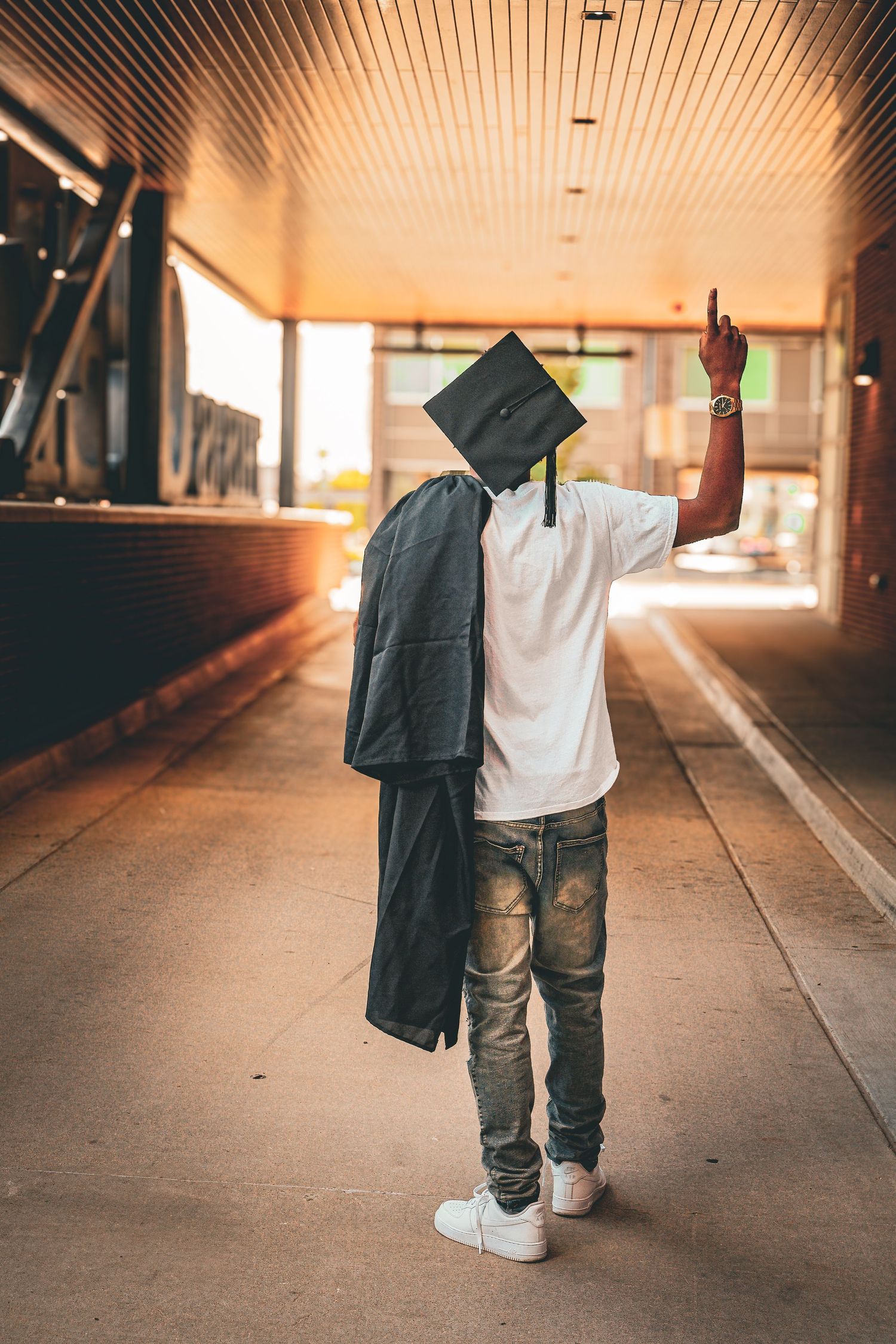 Graduate in cap holding gown over shoulder pointing up in warmly lit corridor.