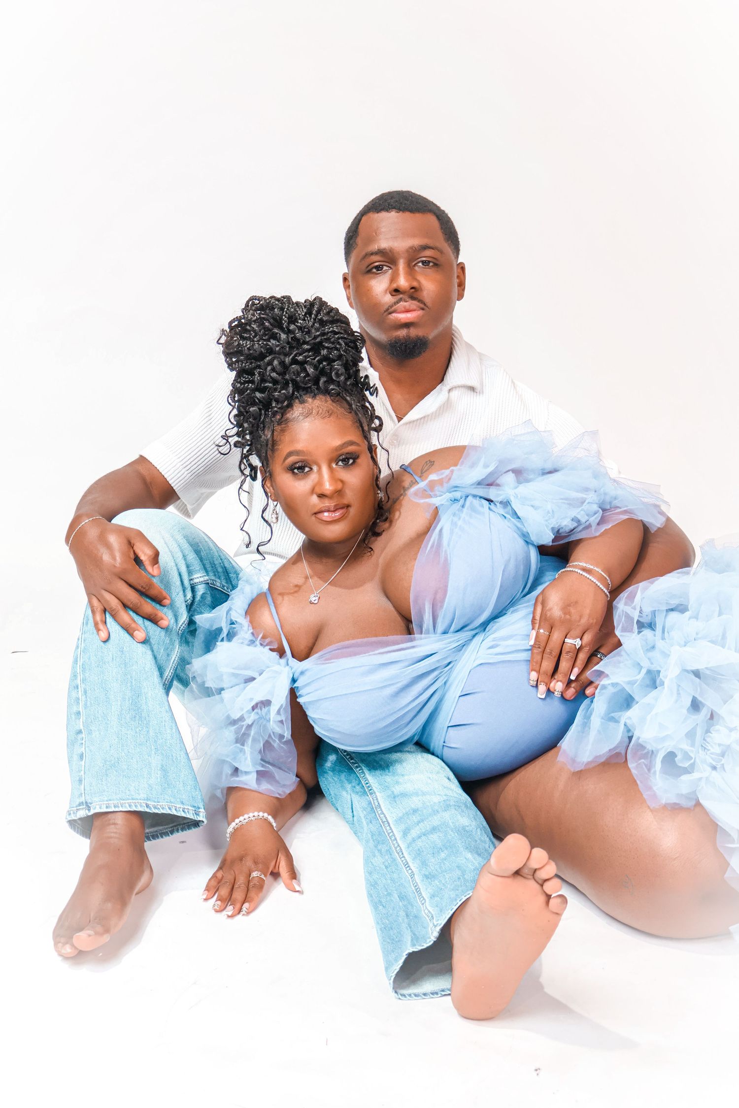 A couple poses together on a white backdrop, woman in blue tulle dress and man in white shirt with jeans.