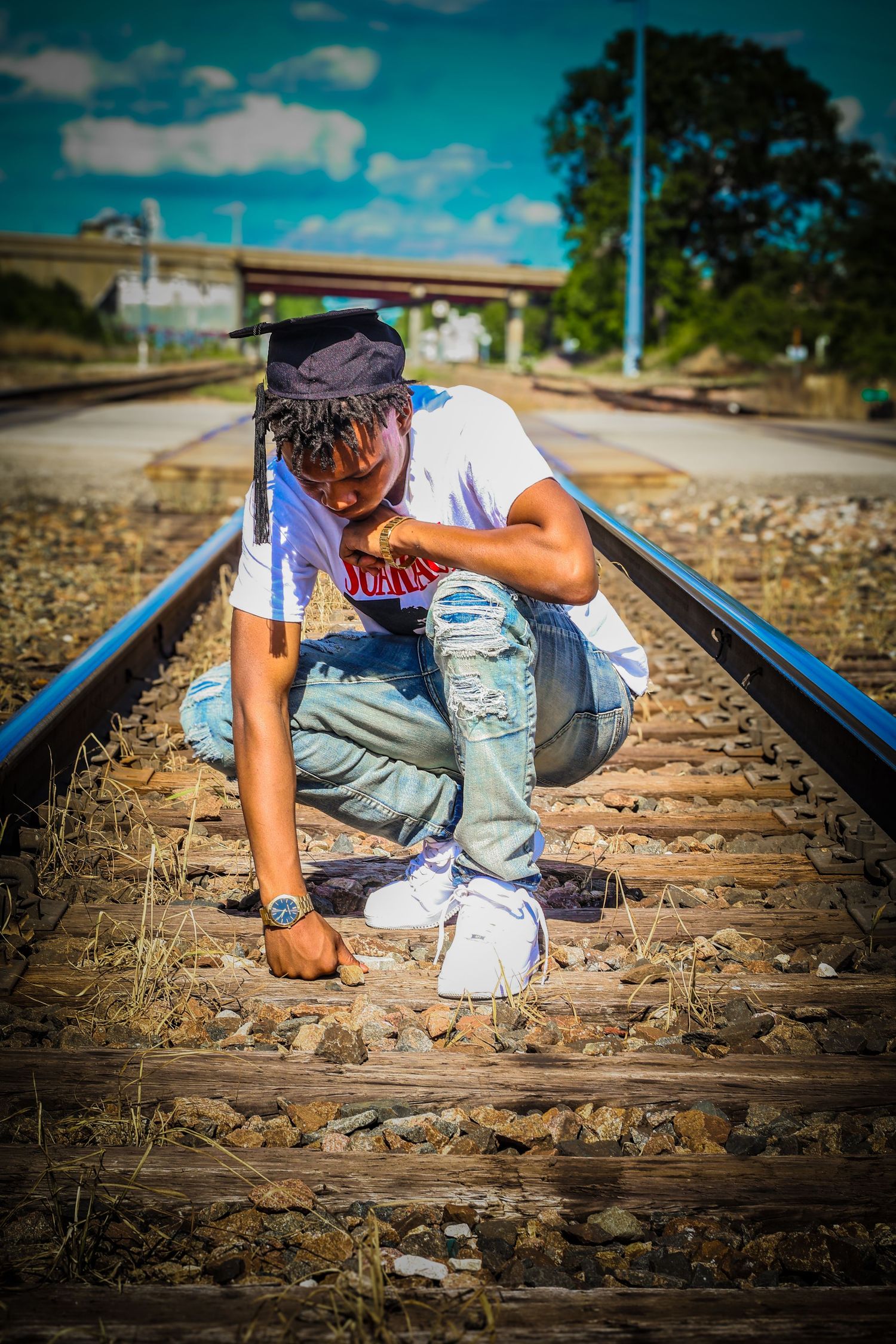 Young man in white shirt and ripped jeans crouching on railway tracks outdoors.