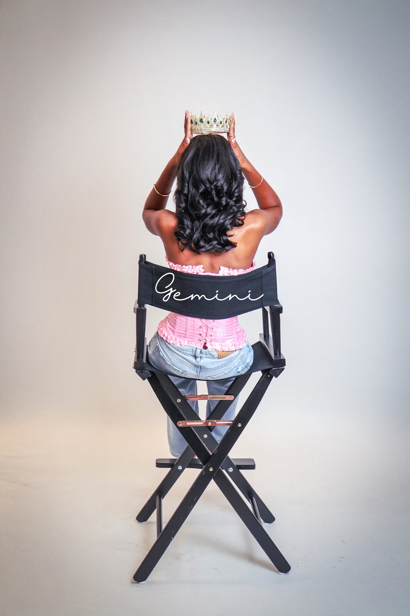Woman sitting in a director's chair labeled Gemini, holding a crown above her head, wearing a pink top and jeans.