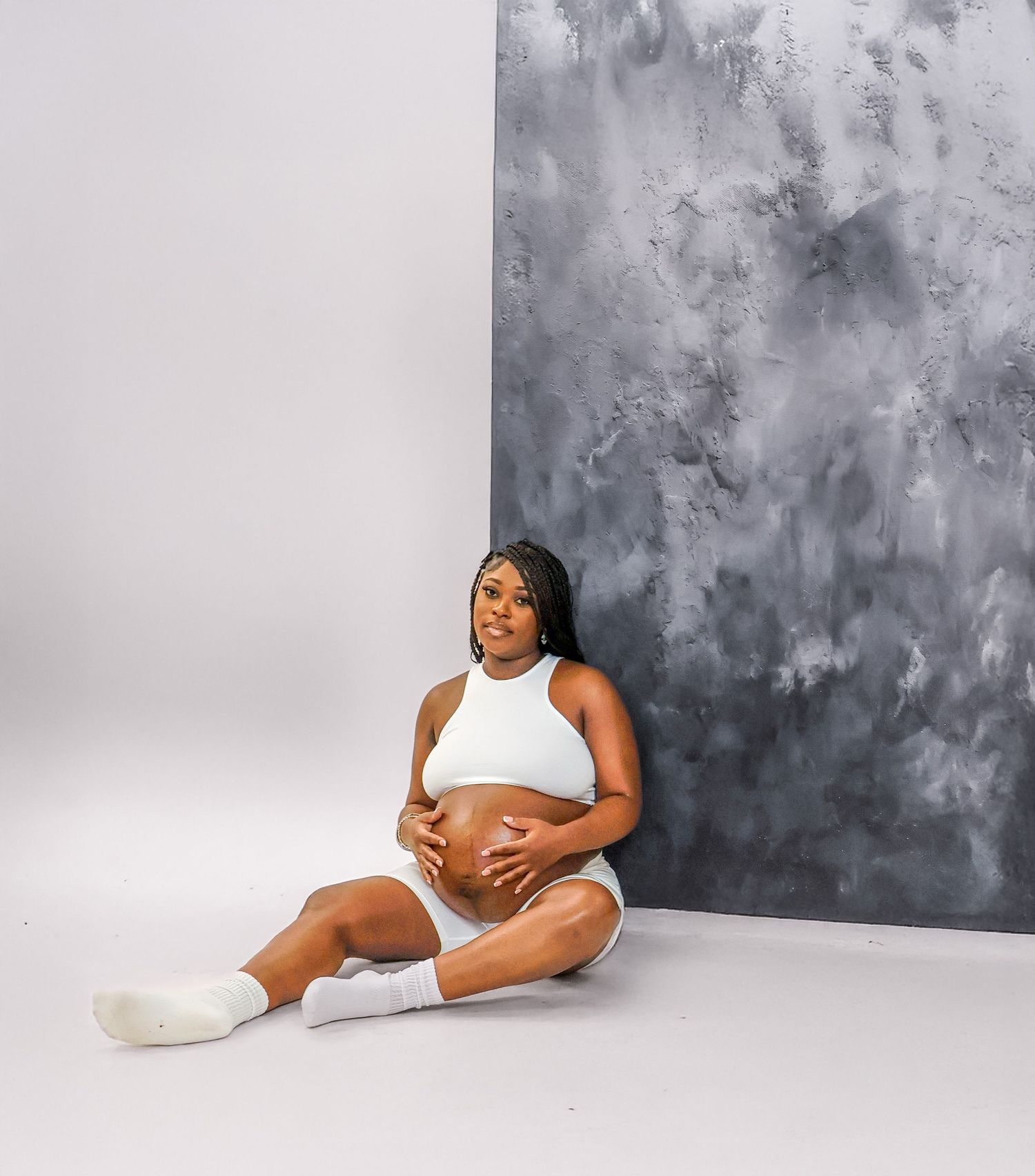 Pregnant woman in white crop top and shorts sitting on studio floor.
