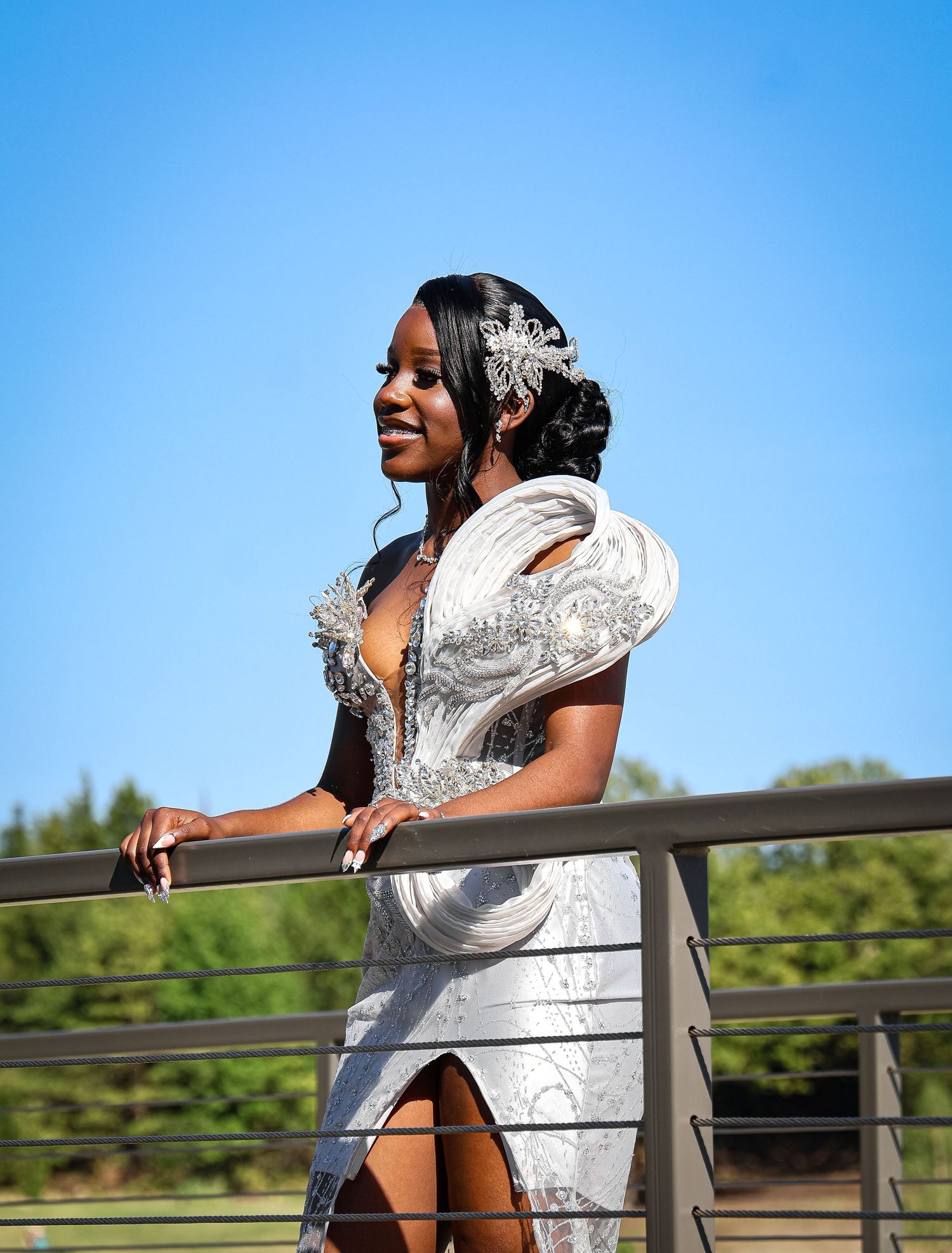 Smiling woman in elegant white beaded gown with crystal headpiece poses gracefully on a railing under a clear blue sky.