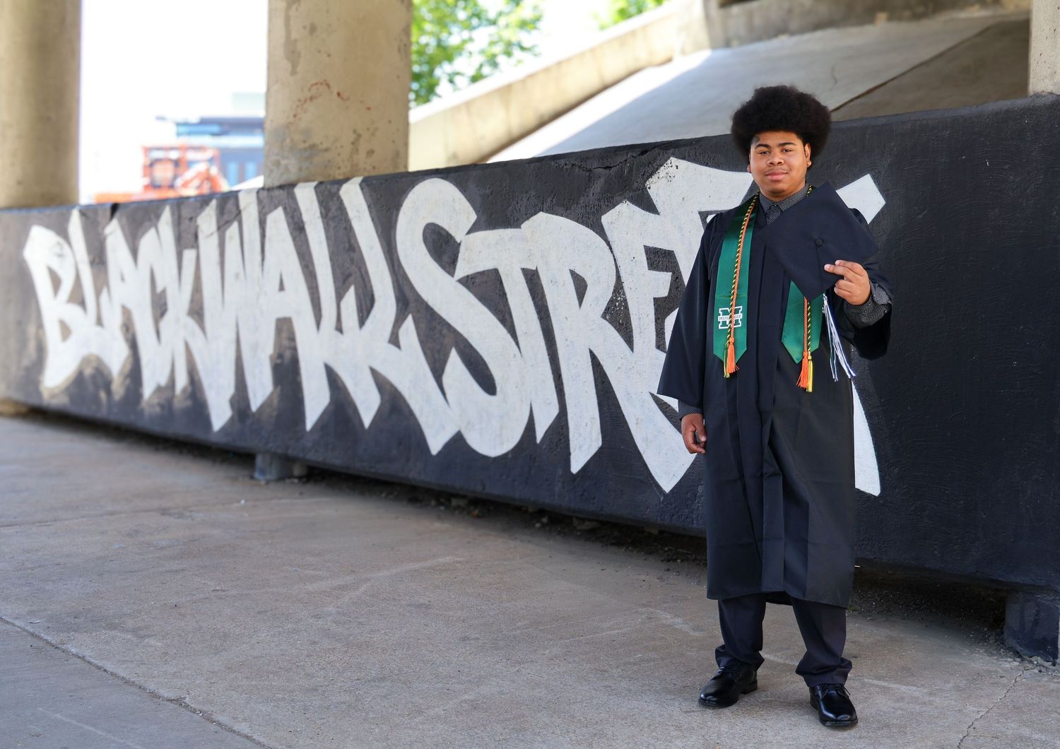 Graduate in cap and gown poses in front of Black Wall Street graffiti mural outdoors.