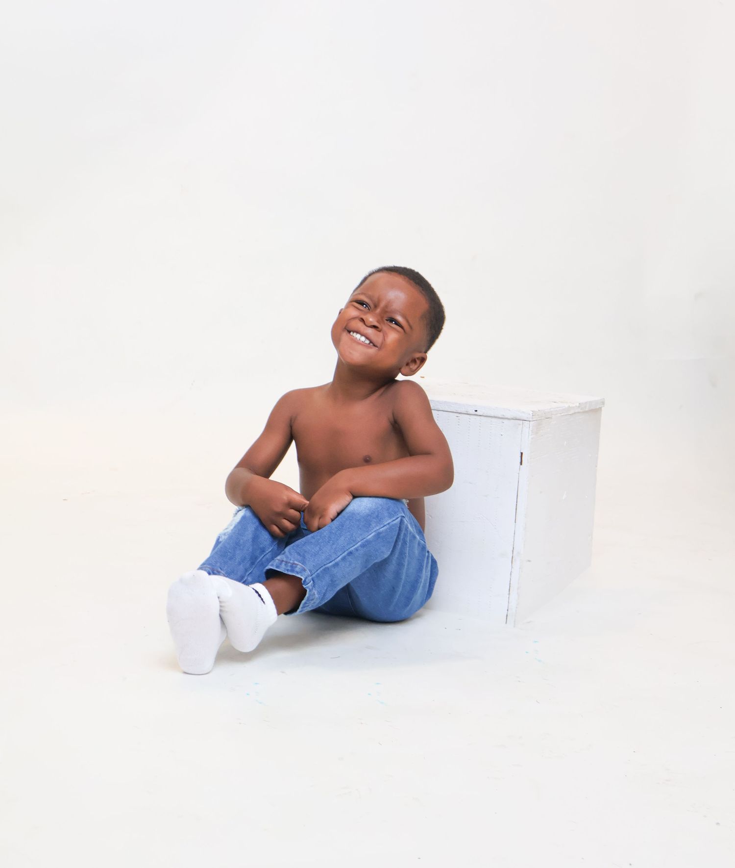 Happy shirtless young boy sitting on floor in jeans and white socks, laughing joyfully in studio.