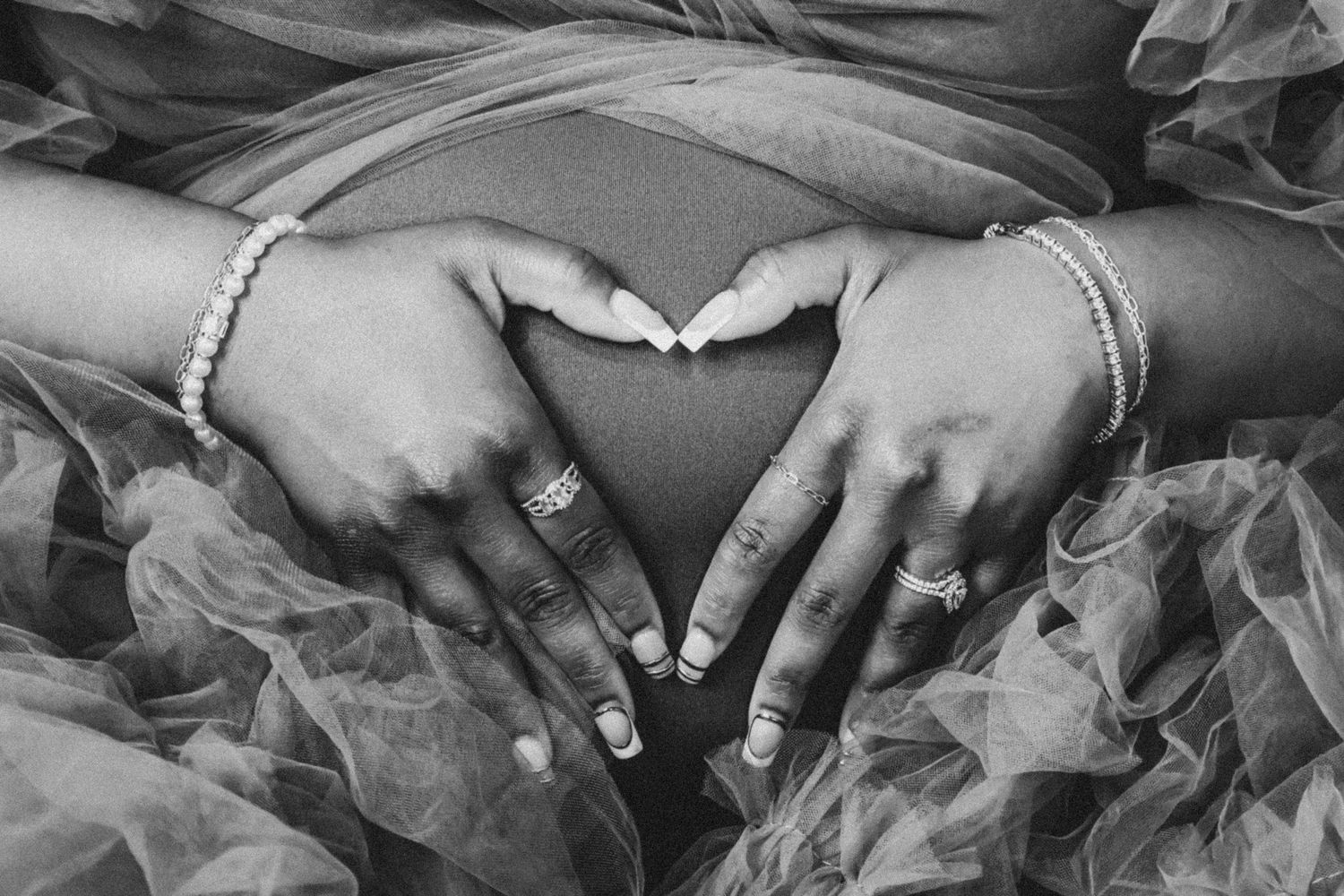 Black and white photo of pregnant woman forming heart shape with hands on baby bump, wearing rings and bracelets.