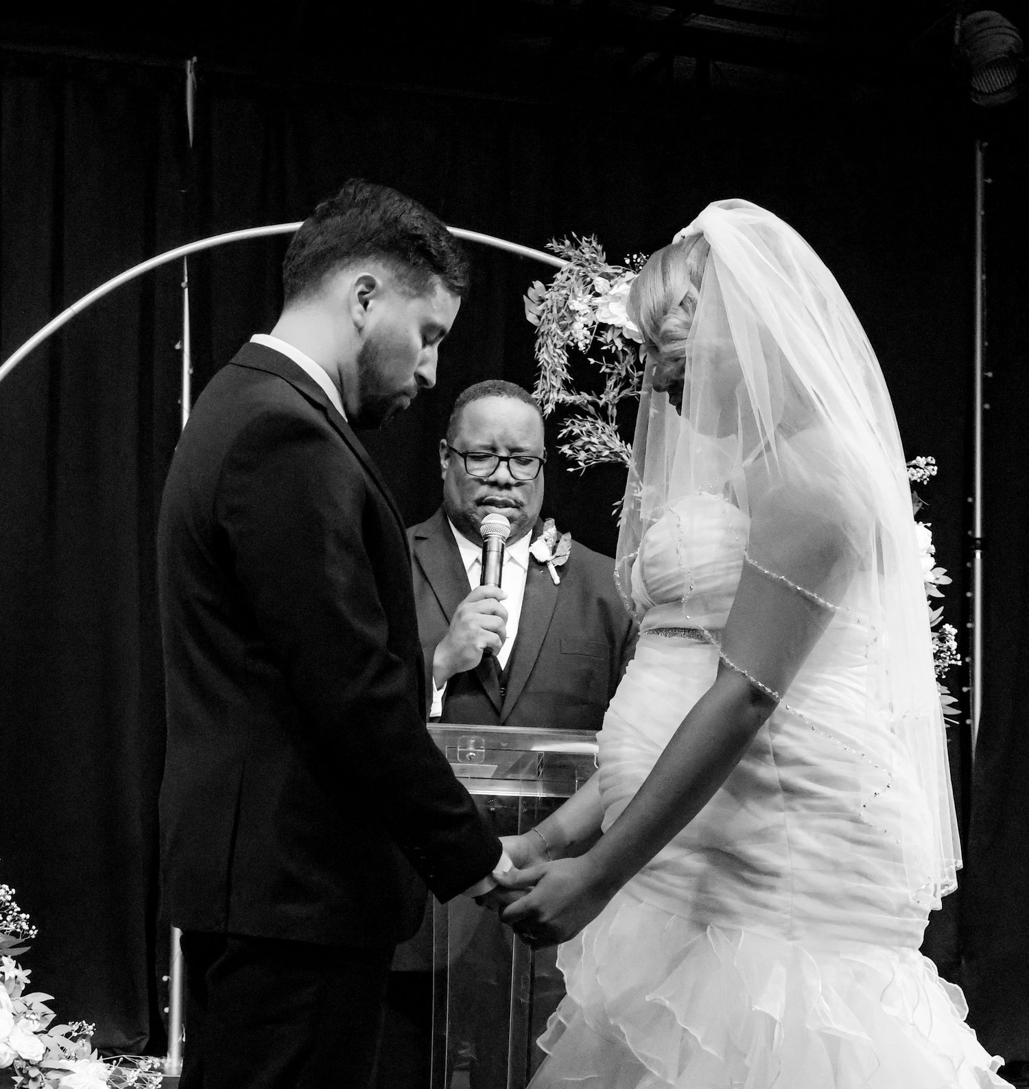 Black and white wedding ceremony photo of bride and groom holding hands before an officiant at the altar.