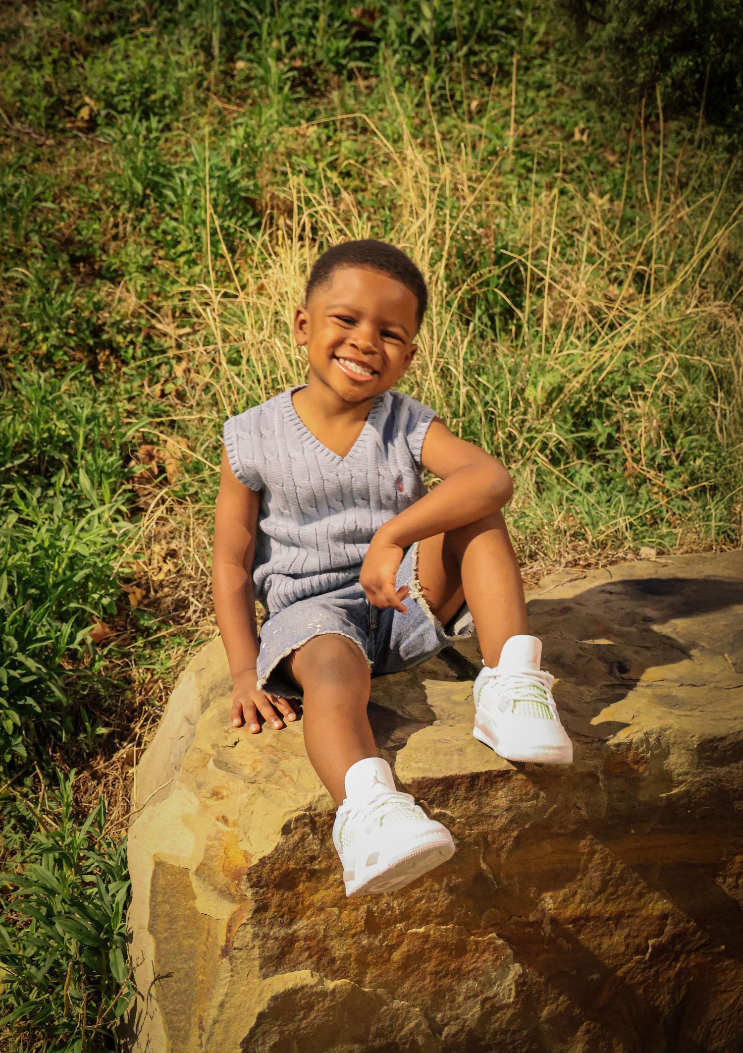 Happy young boy smiling while sitting on a rock outdoors in a grey vest and shorts.