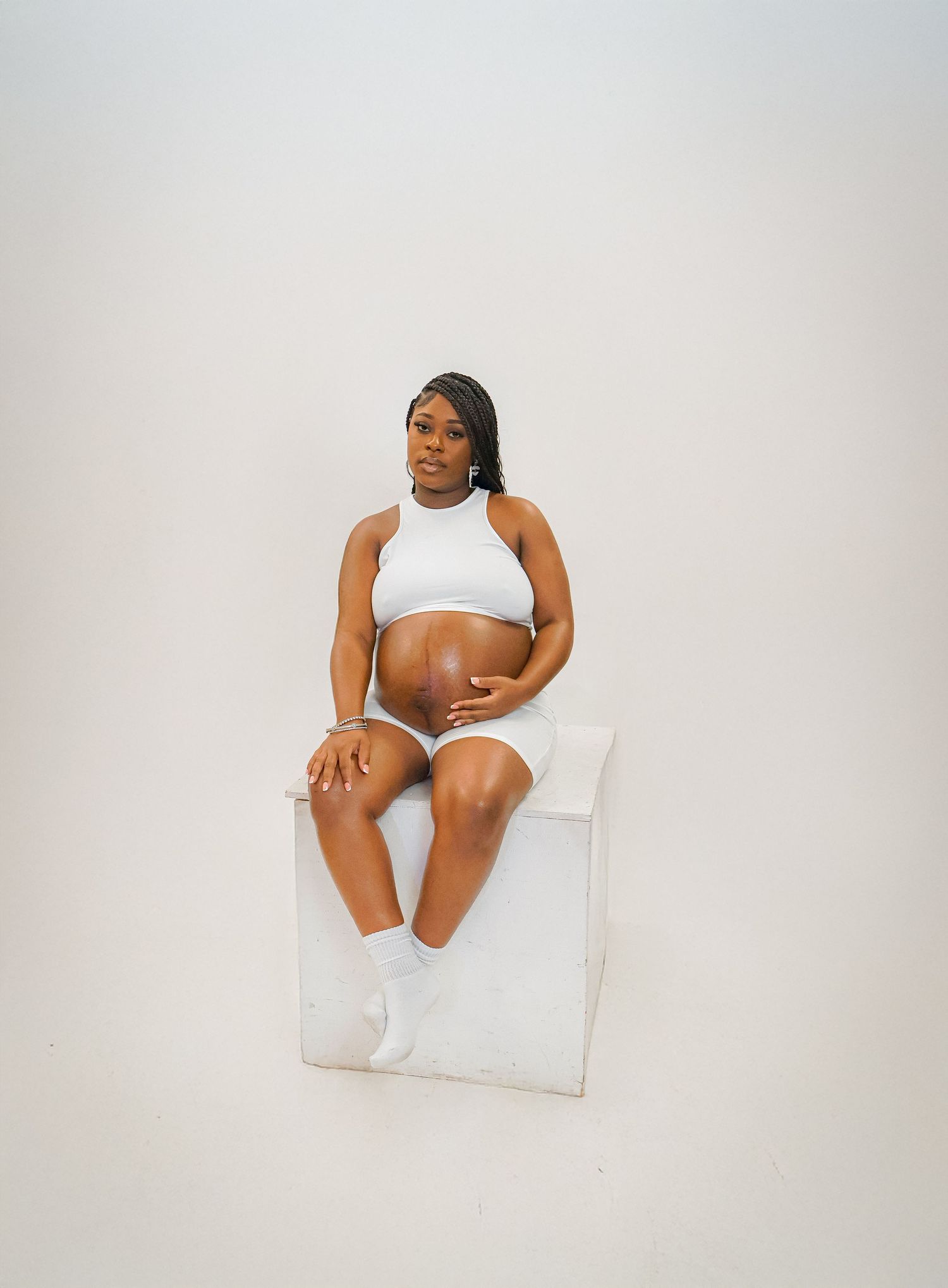 Pregnant woman in white crop top and shorts sitting on white cube, cradling baby bump in a bright studio.