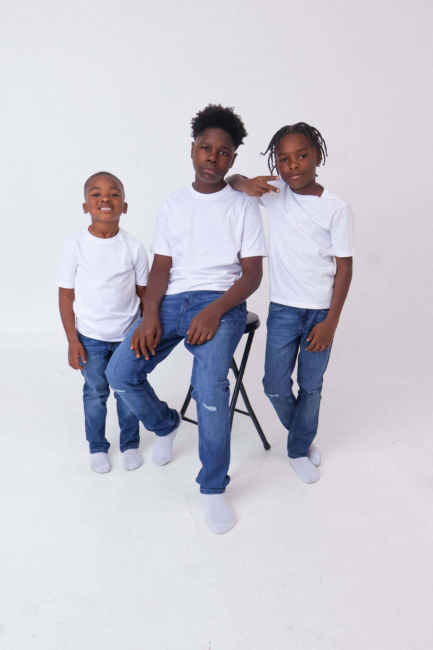 Three young boys in white t-shirts and jeans pose together in a bright white photography studio.