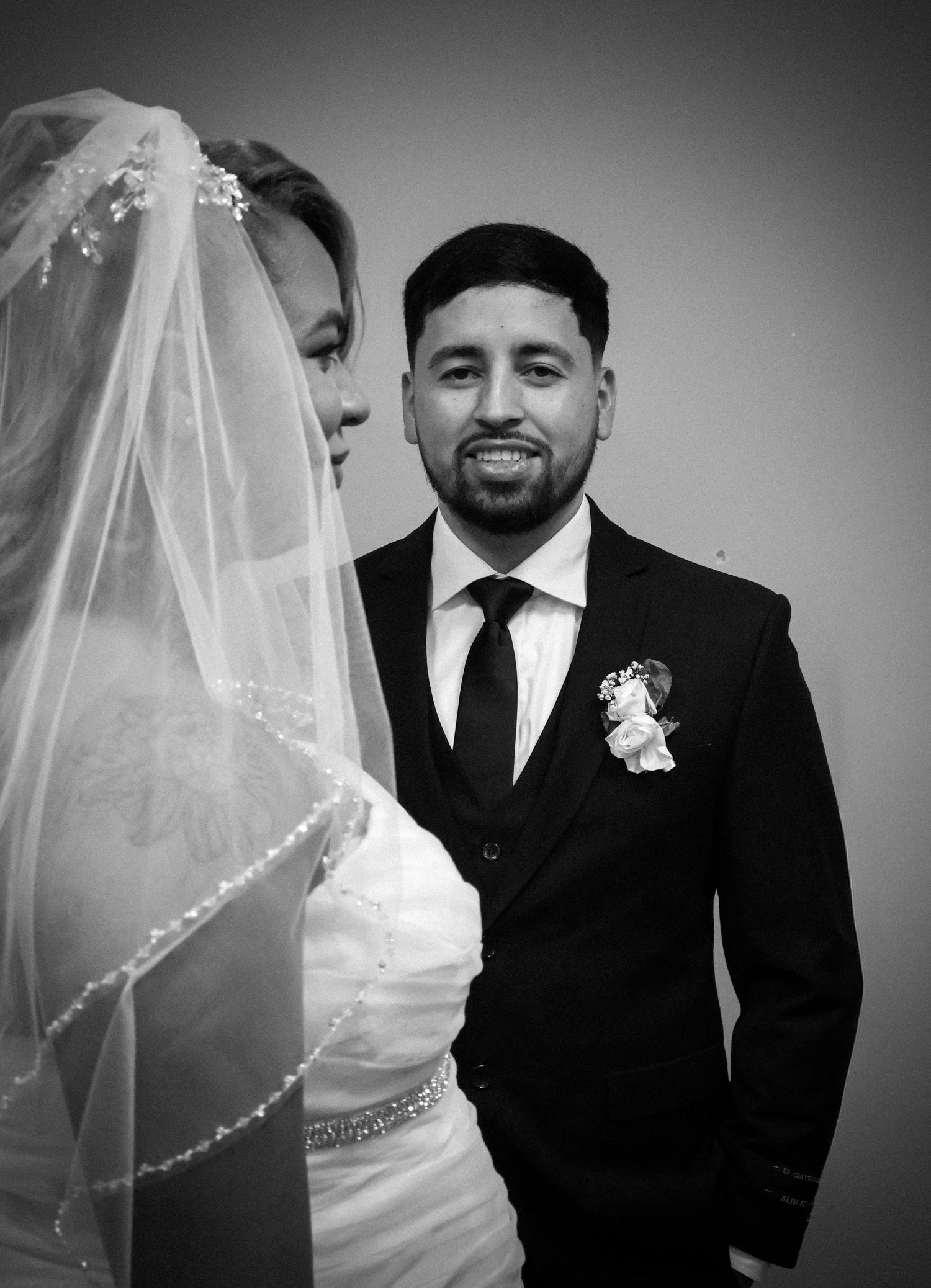 Groom in black suit smiling beside veiled bride in black and white wedding portrait.