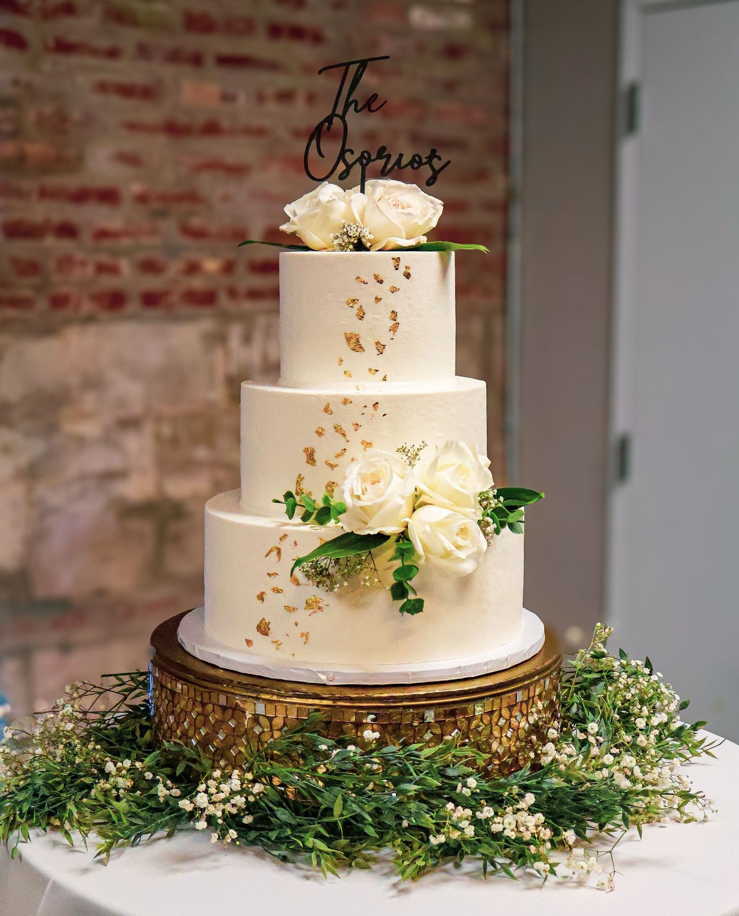 Elegant four-tier white wedding cake with gold leaf, white roses, greenery, and The Osarios cake topper on a gold stand.