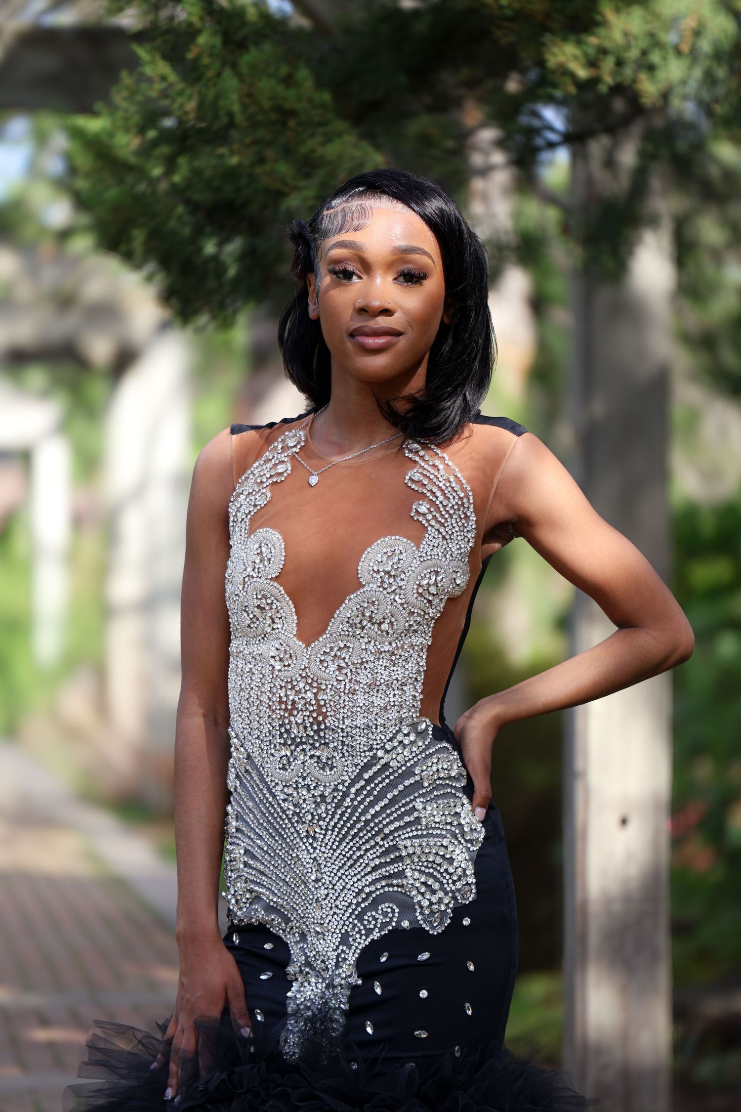 Young woman in elegant black and silver beaded mermaid gown poses outdoors among lush greenery.