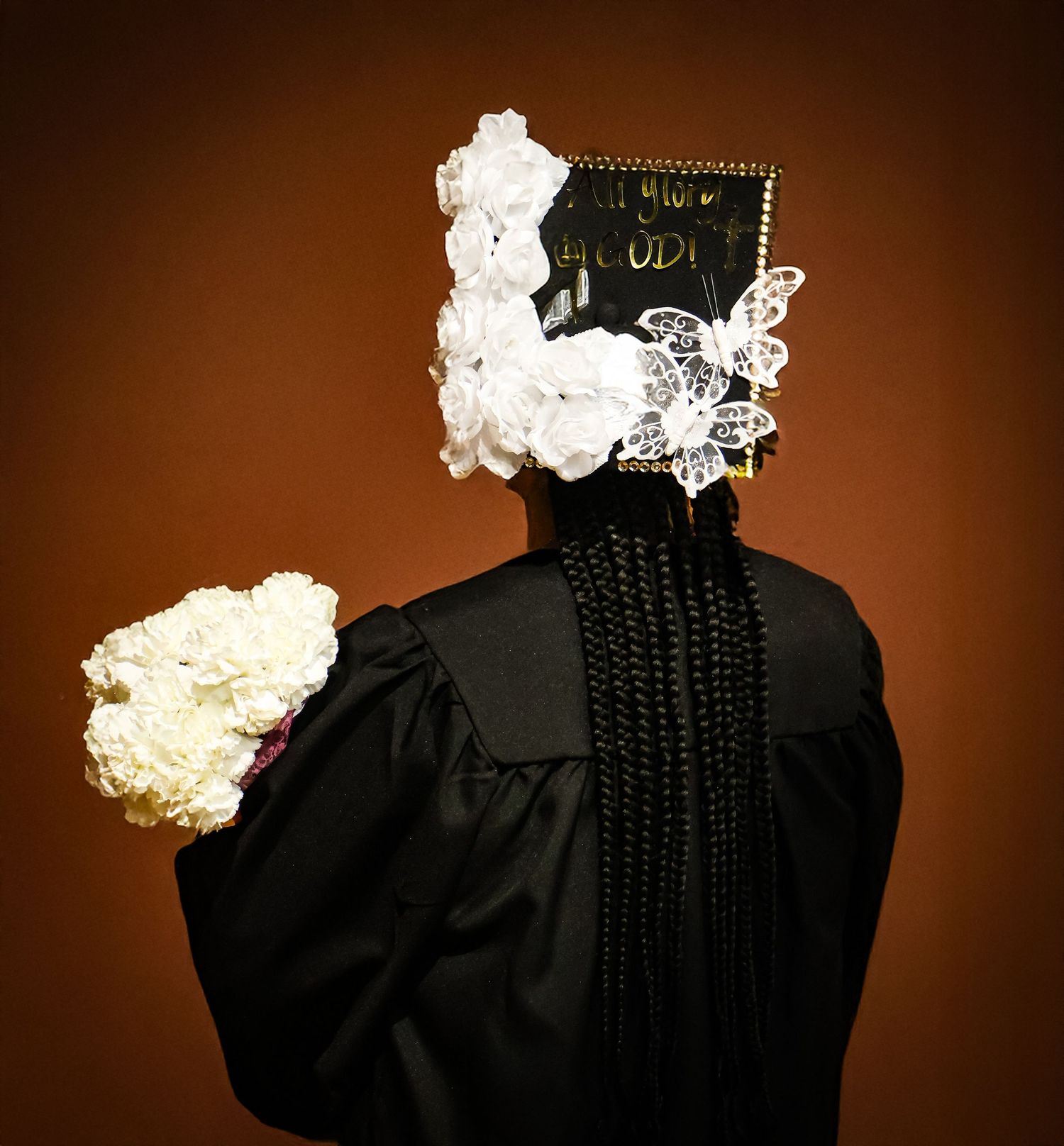 Back view of graduate in black gown with decorated floral mortarboard cap holding white flower bouquet.