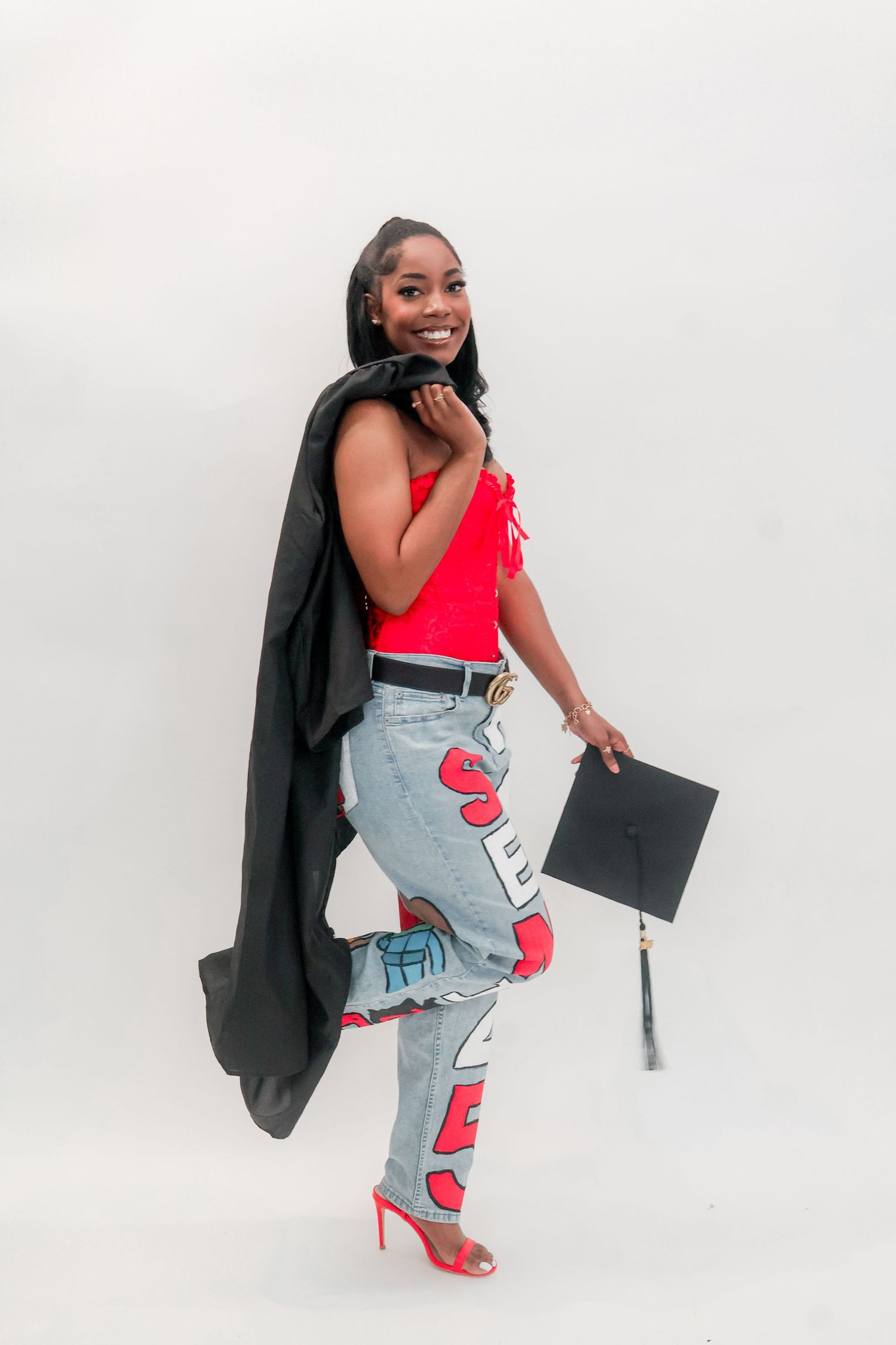 Graduate smiles over shoulder in red top, custom jeans, holding black cap with gown draped over arm.
