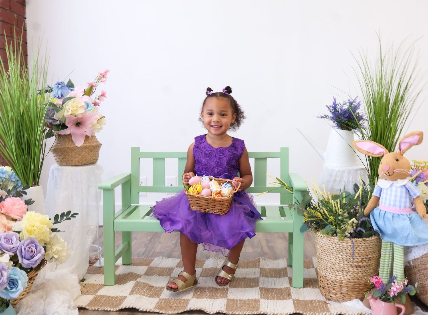 Adorable young girl in purple dress sitting on green bench holding Easter basket with colorful eggs.