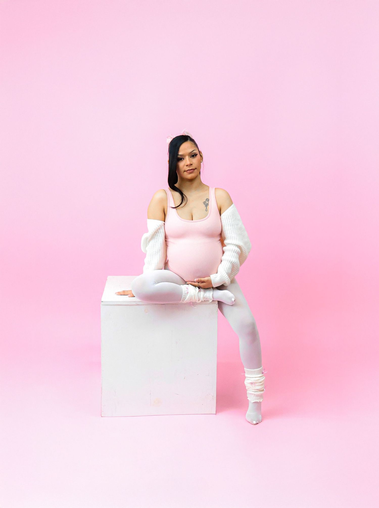 Pregnant woman in white outfit sitting on cube against pink background, cradling baby bump in maternity photoshoot.