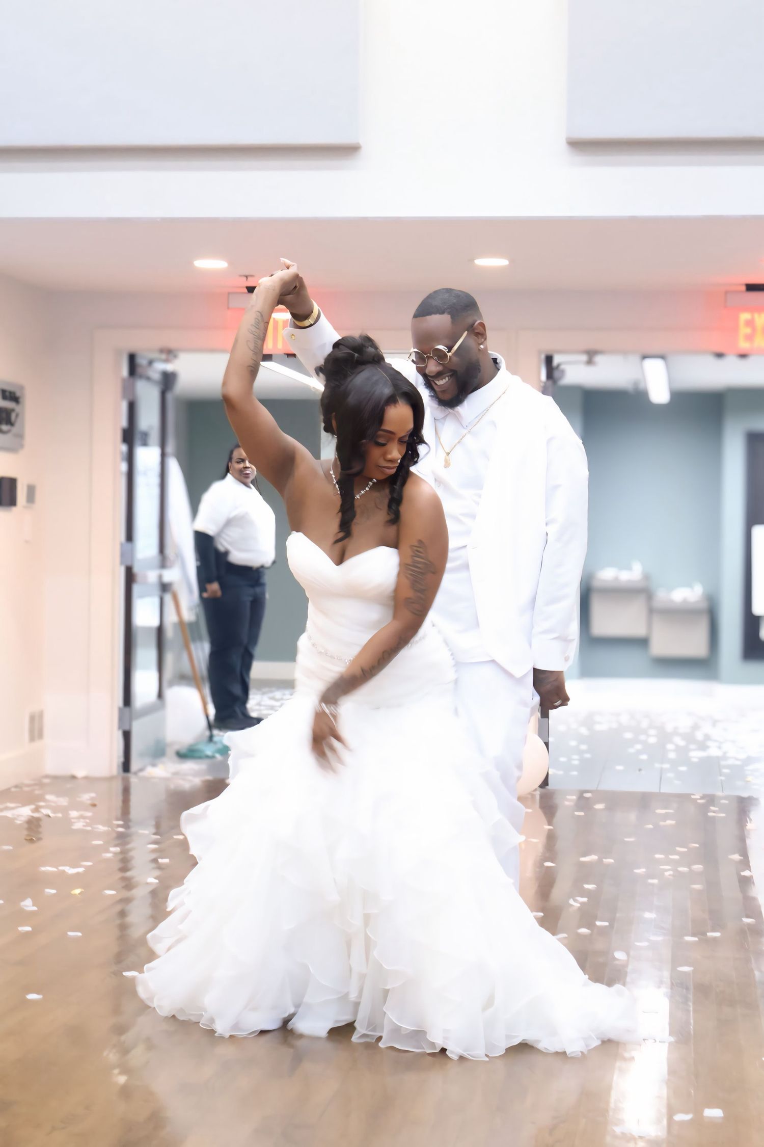 Bride in ruffled white gown twirls during first dance with groom in white suit at reception.
