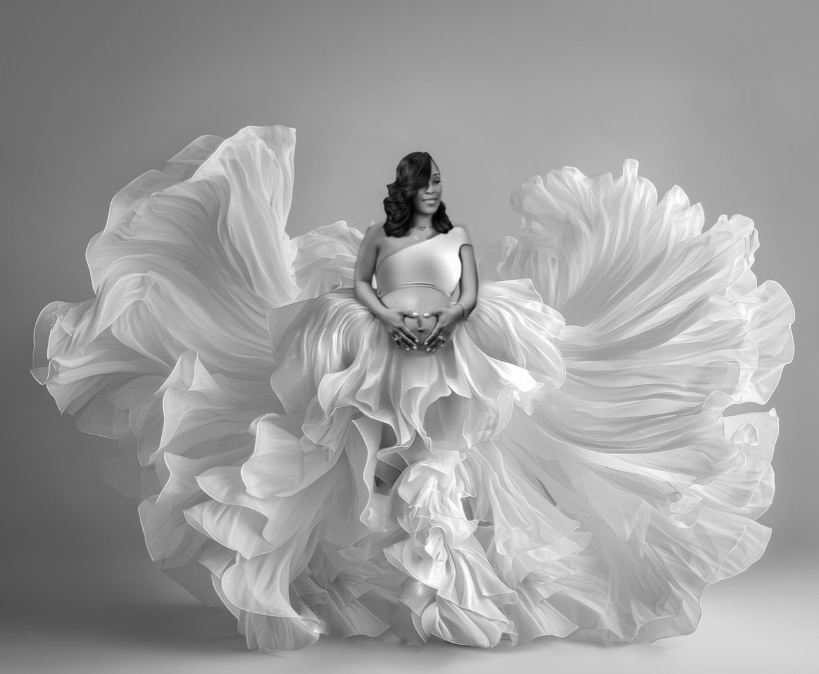 A pregnant woman in a flowing white gown poses gracefully in a dramatic black and white studio photo.