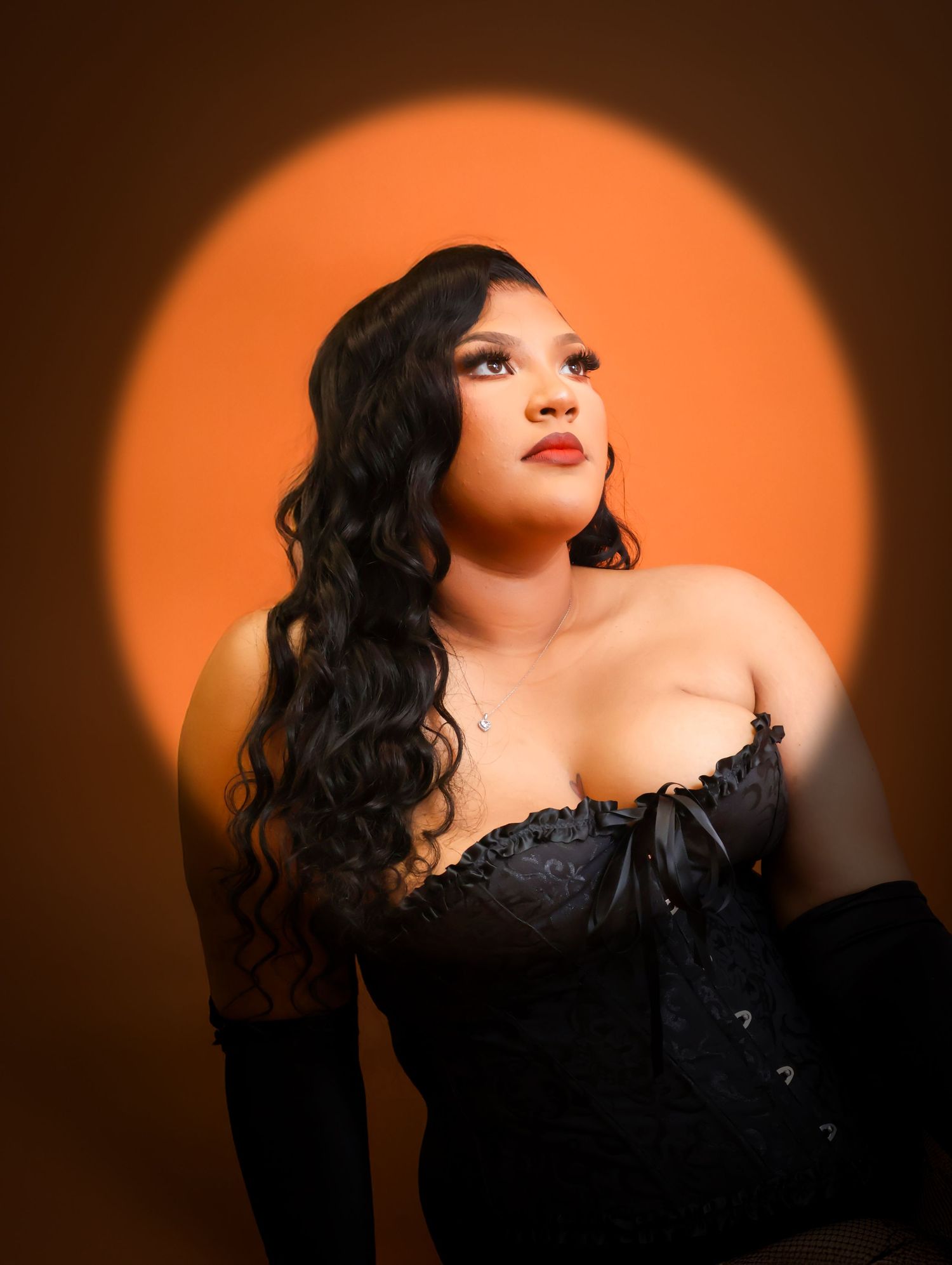 Young woman in black lace corset and gloves posing dramatically against warm orange spotlight background.