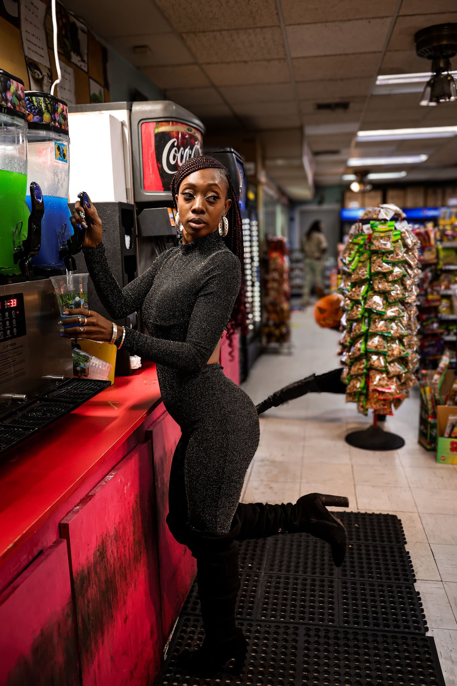 A woman in a black catsuit poses playfully at a Coca-Cola fountain inside a convenience store.