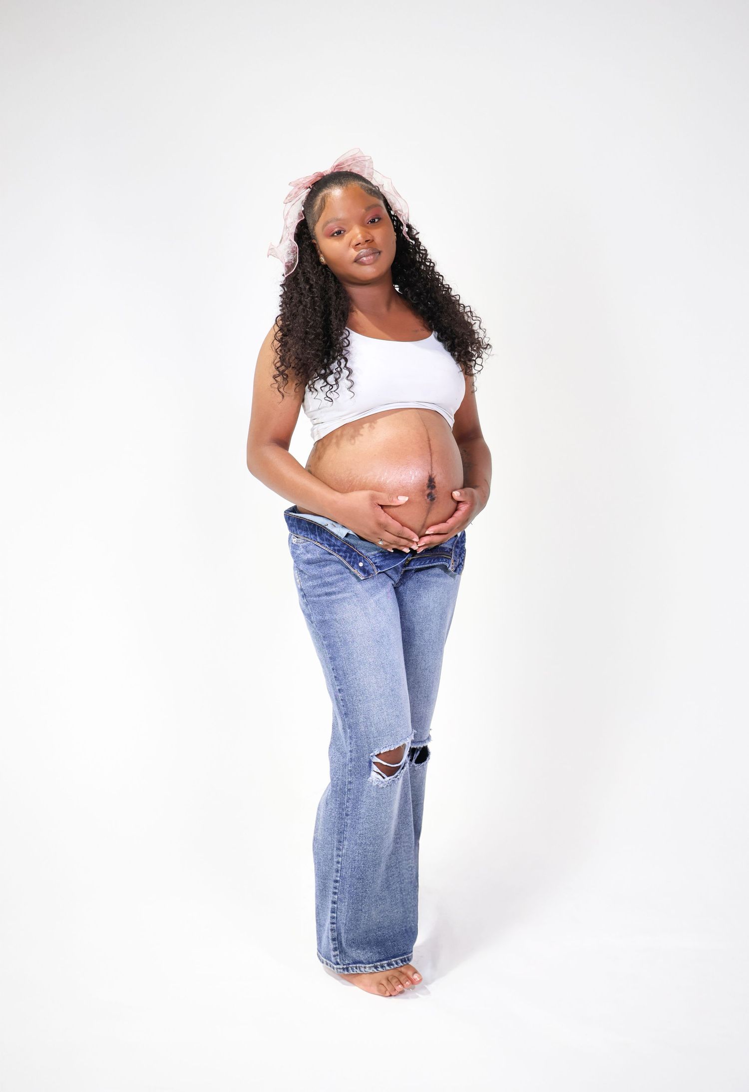 Pregnant woman in white crop top and jeans cradling baby bump, wearing pink bow headband, studio photo.