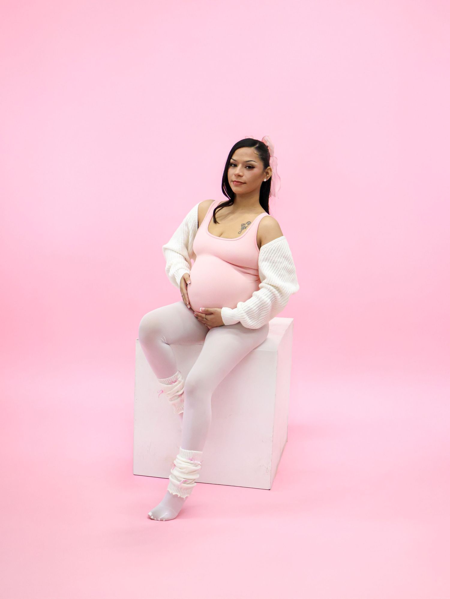 Pregnant woman in pink bodysuit, white leggings and cardigan poses on white cube against pink background.