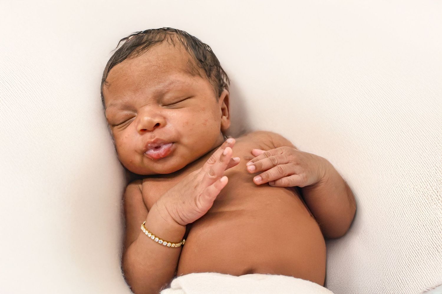 Peaceful sleeping newborn baby wearing a gold bracelet, lying on a white blanket.