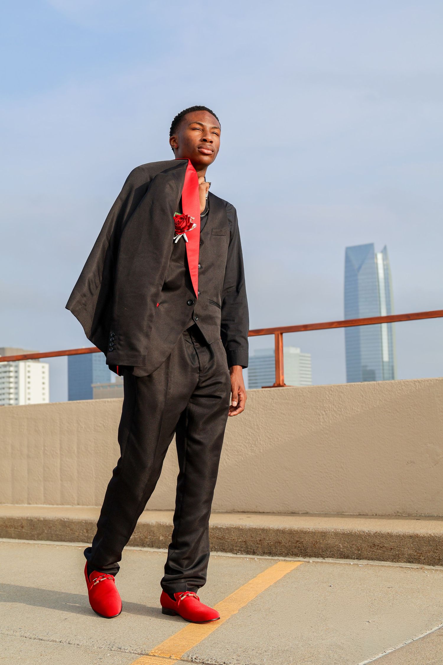 Young Black man in black suit with red tie and red shoes posing on rooftop with city skyline background.