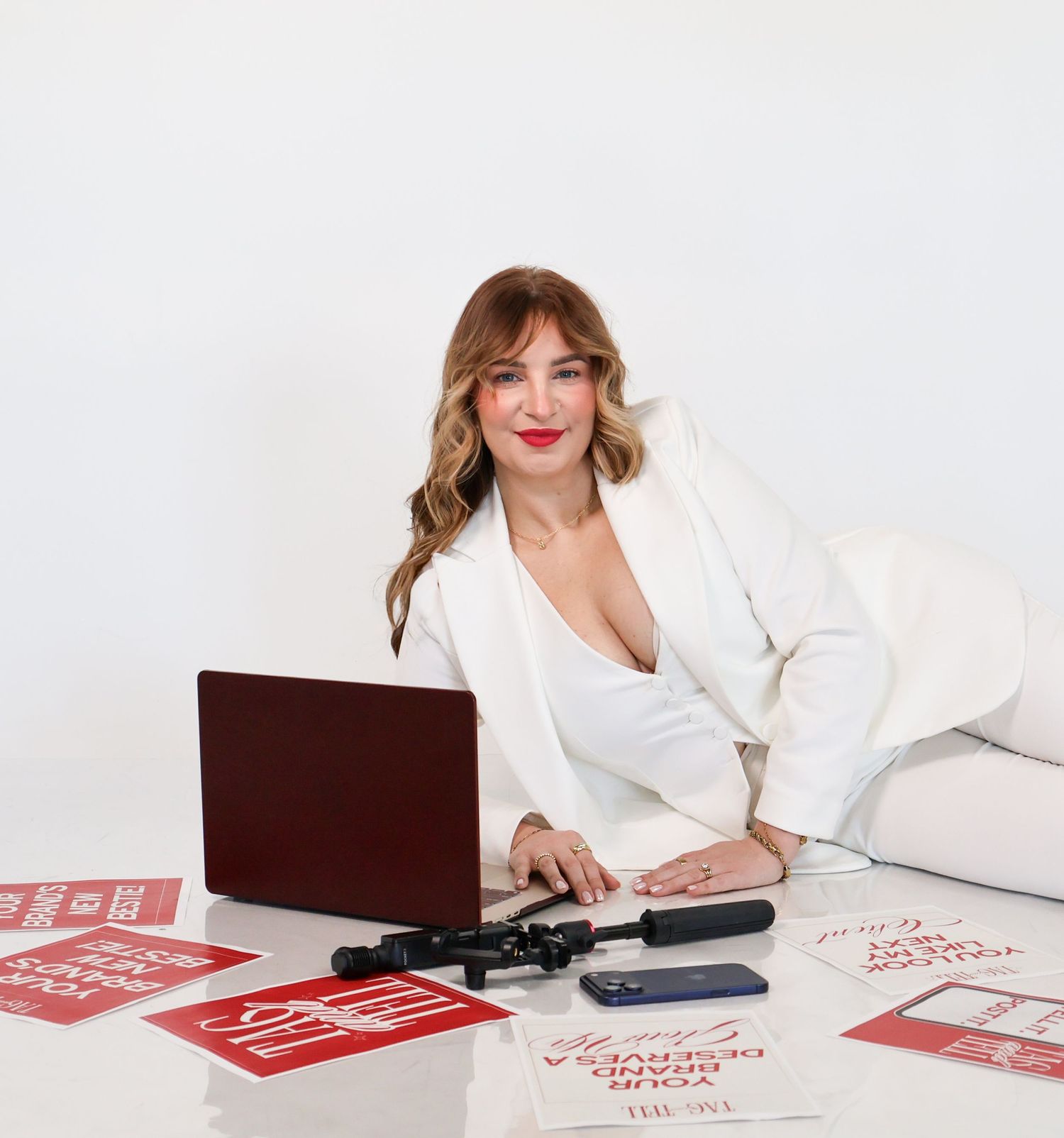 Woman in white blazer sits at desk with laptop, red marketing signs, and beauty tools in a bright studio.
