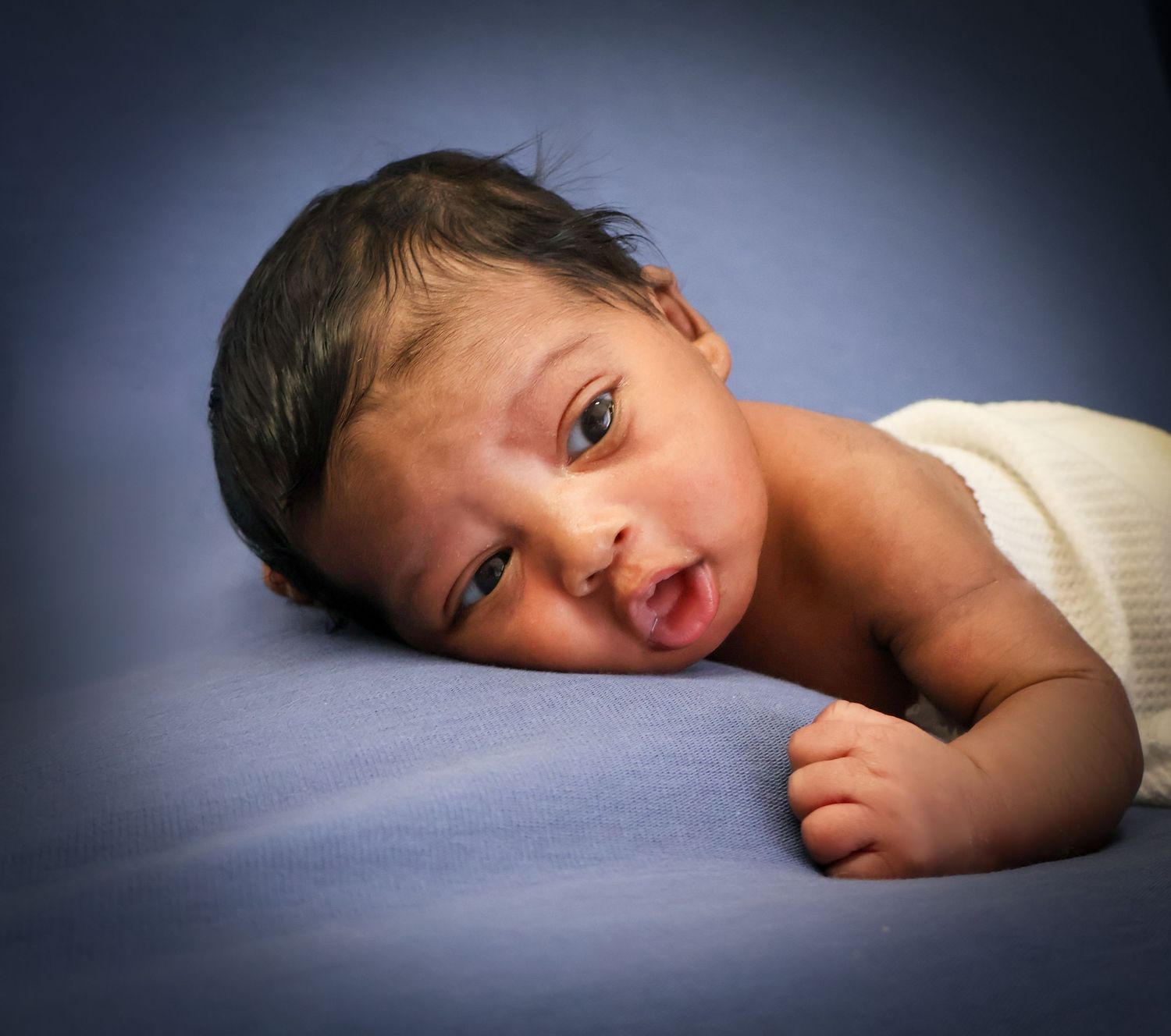 Newborn baby with dark hair lying on a blue blanket, looking up with wide eyes and open mouth.