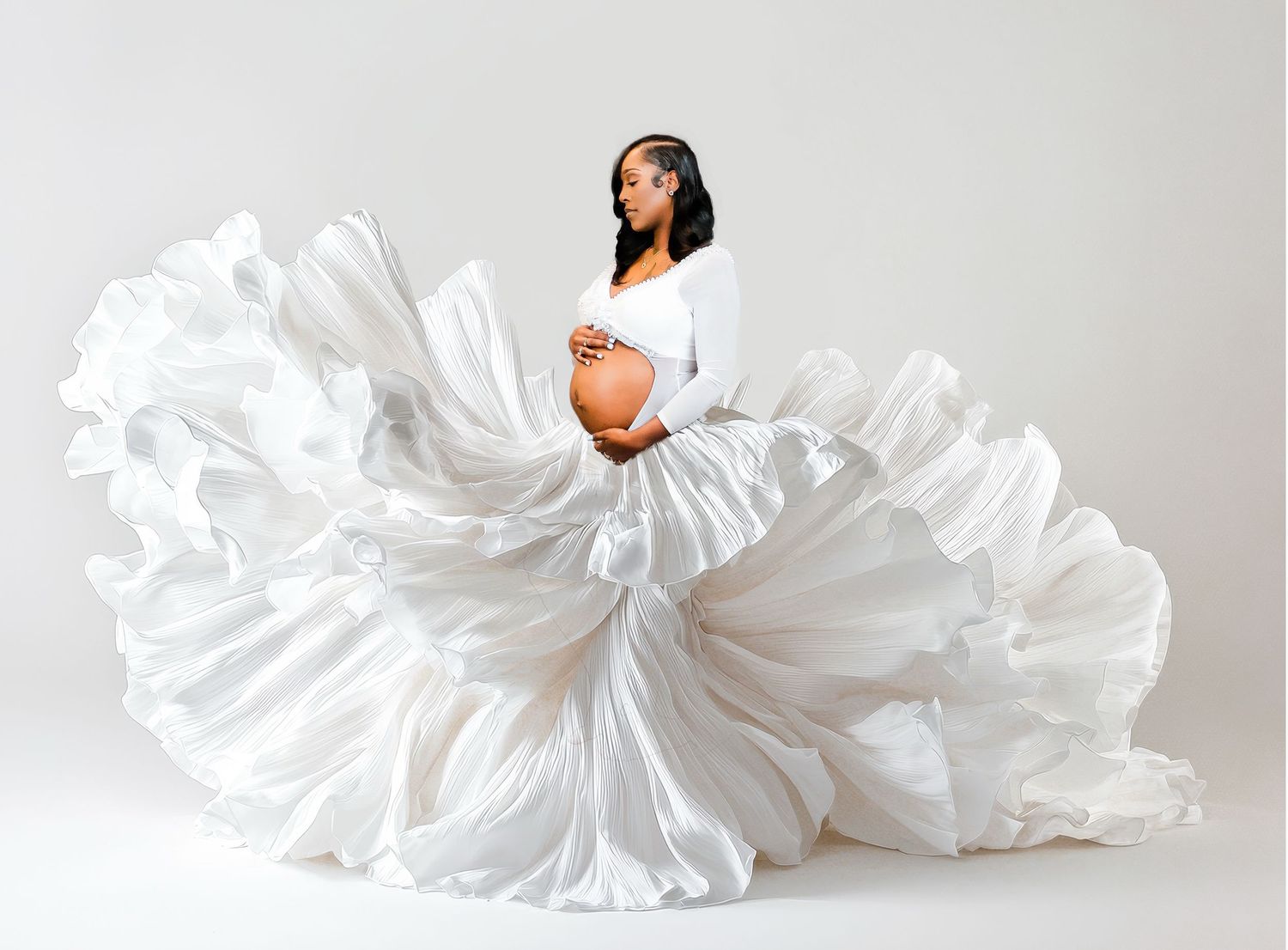 Pregnant woman in flowing white satin gown holding baby bump in elegant maternity photo studio session.