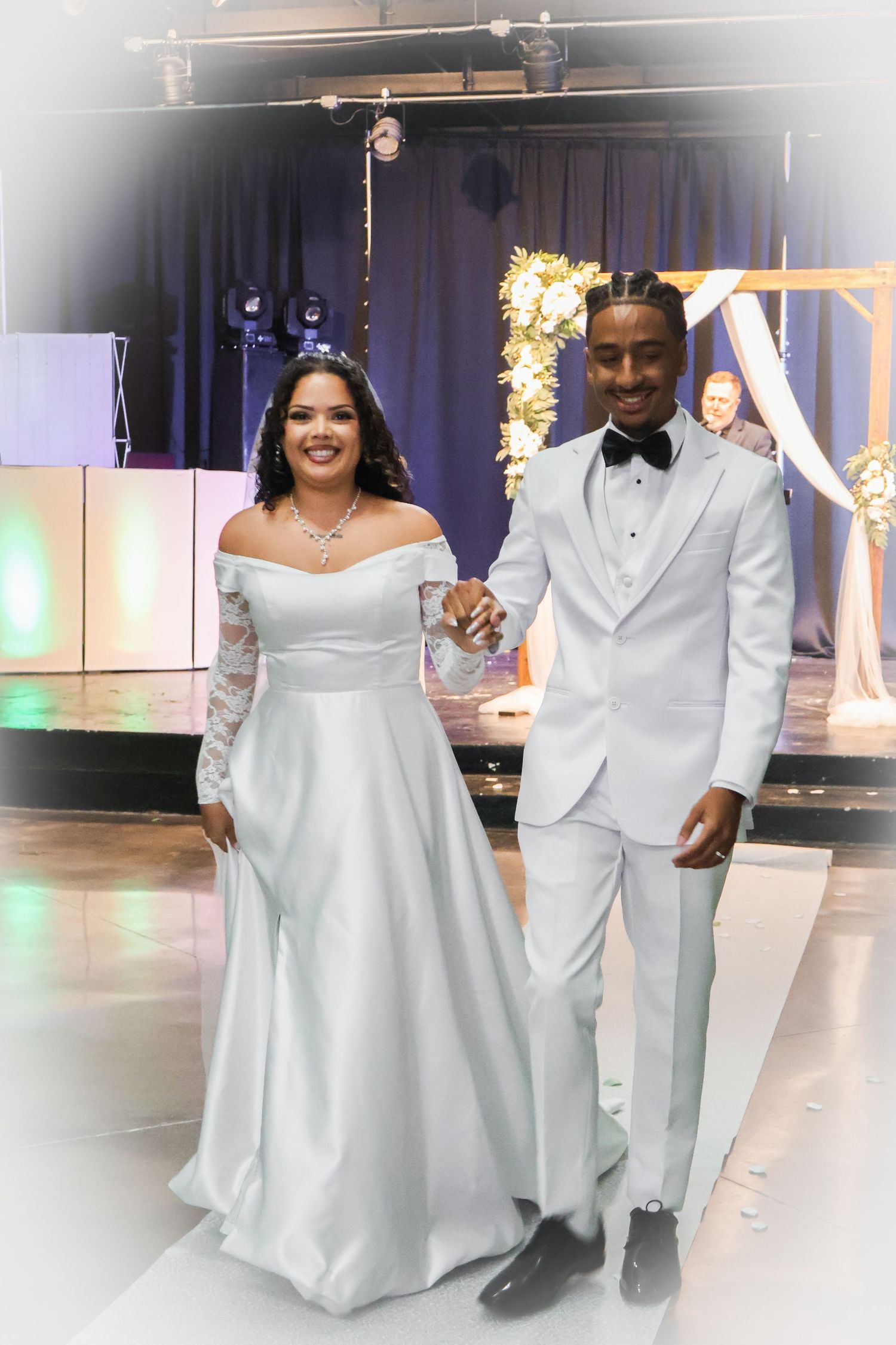 Bride in white off-shoulder gown and groom in white tuxedo holding hands at elegant wedding reception.