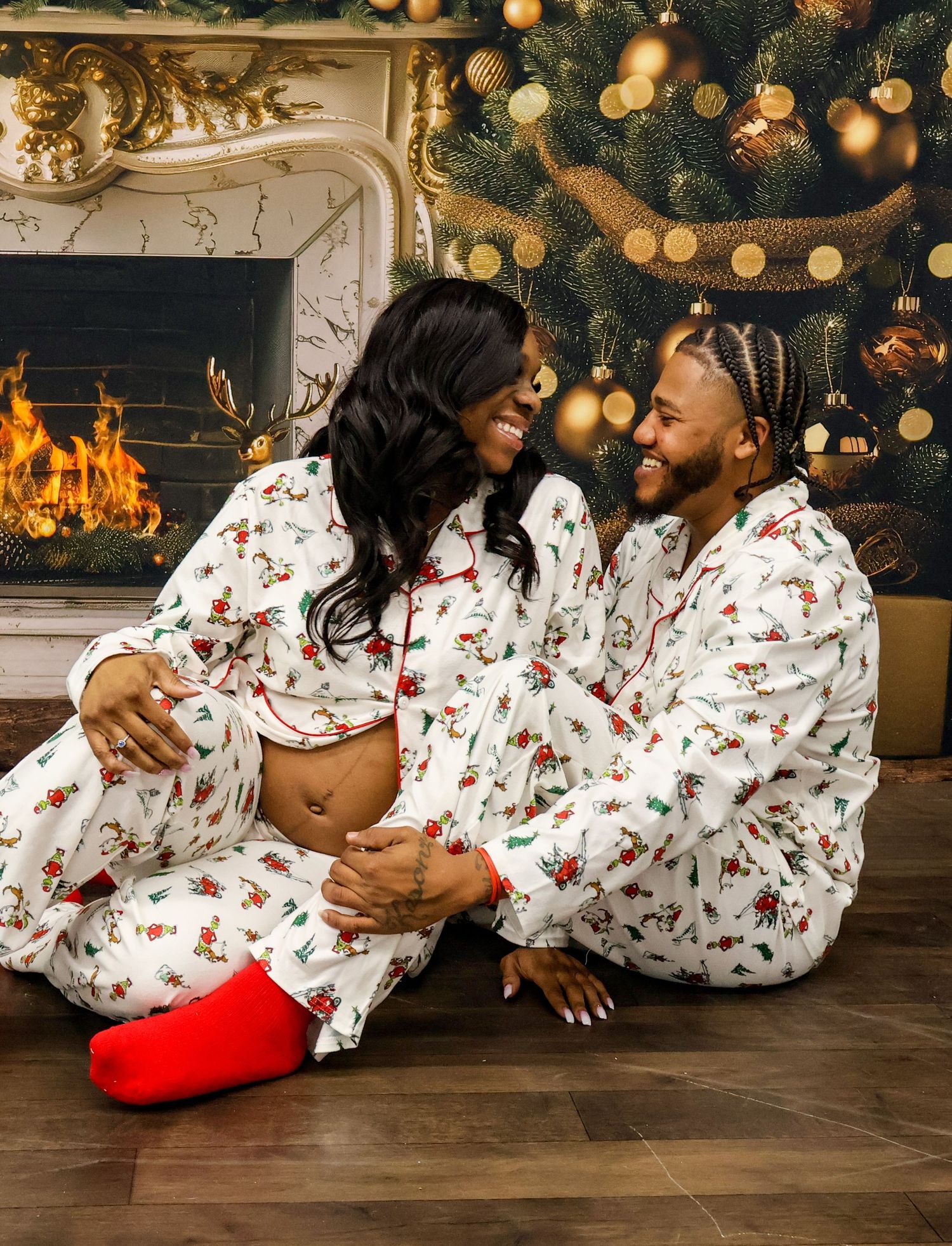 Couple in matching Christmas pajamas sitting by fireplace and decorated tree, woman showing baby bump.