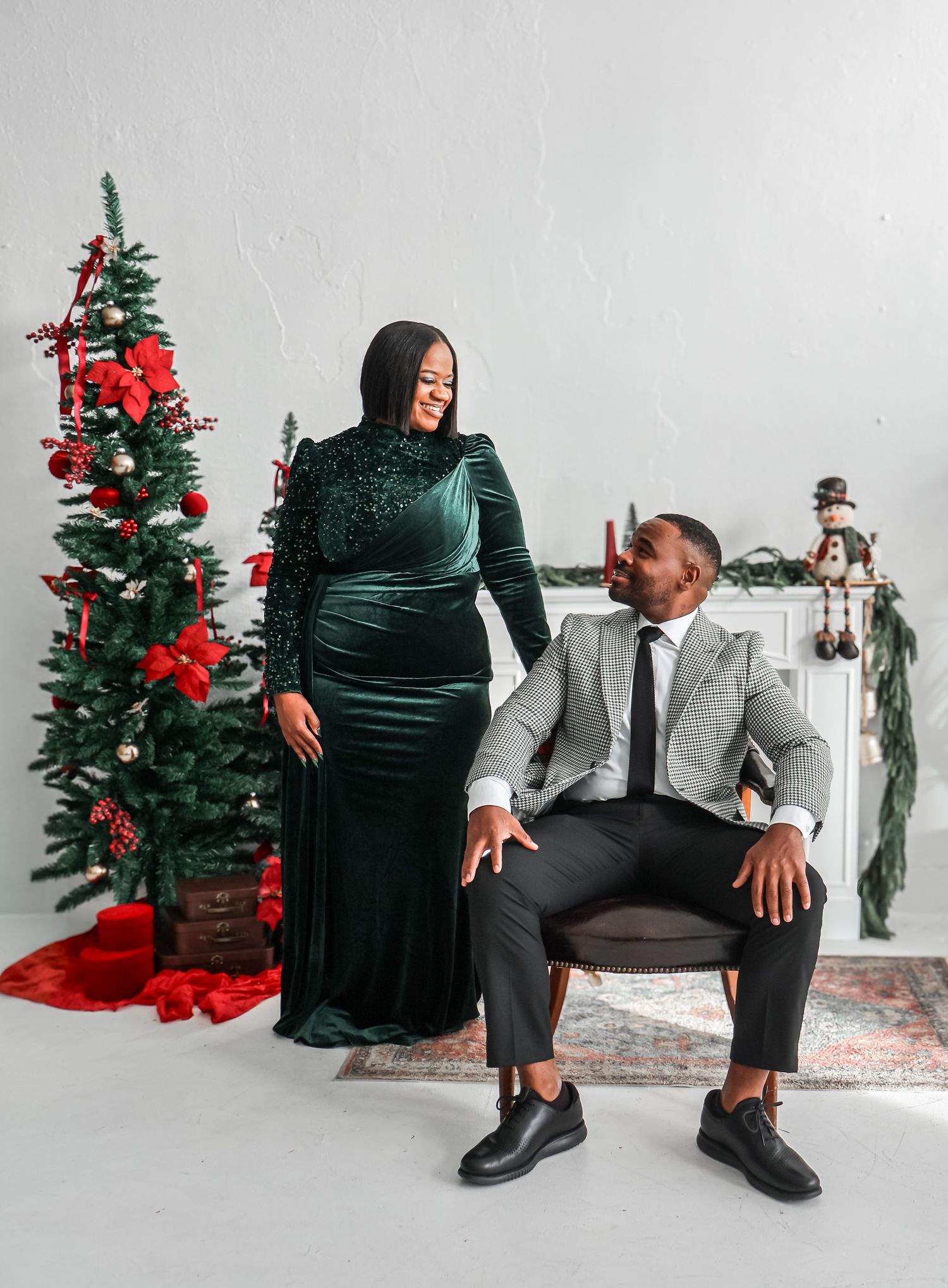 Couple in elegant Christmas photo shoot, woman in green velvet gown, man in gray suit, beside decorated holiday tree.