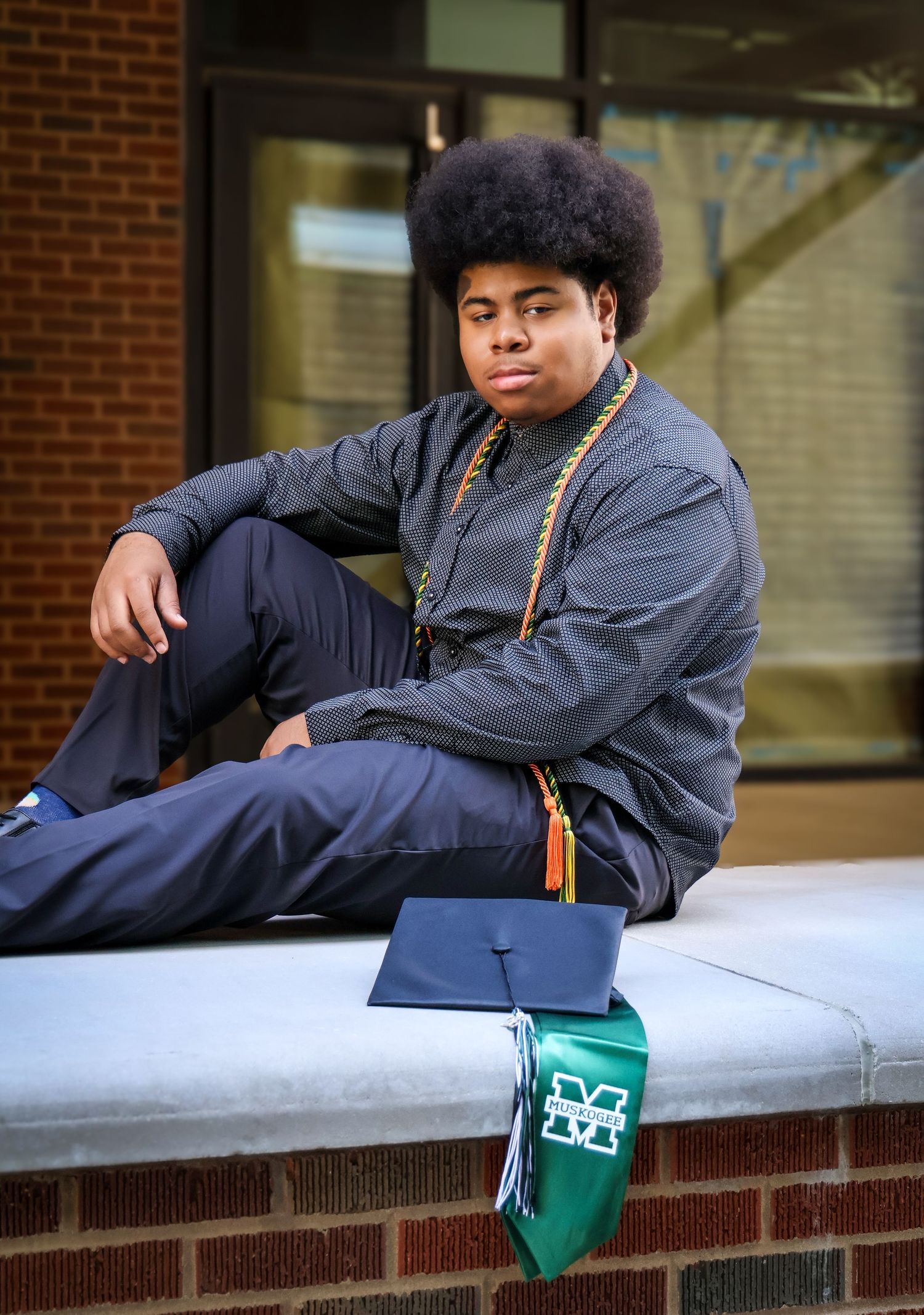 Young man in graduation attire with afro hairstyle sitting on brick wall with cap and green stole.