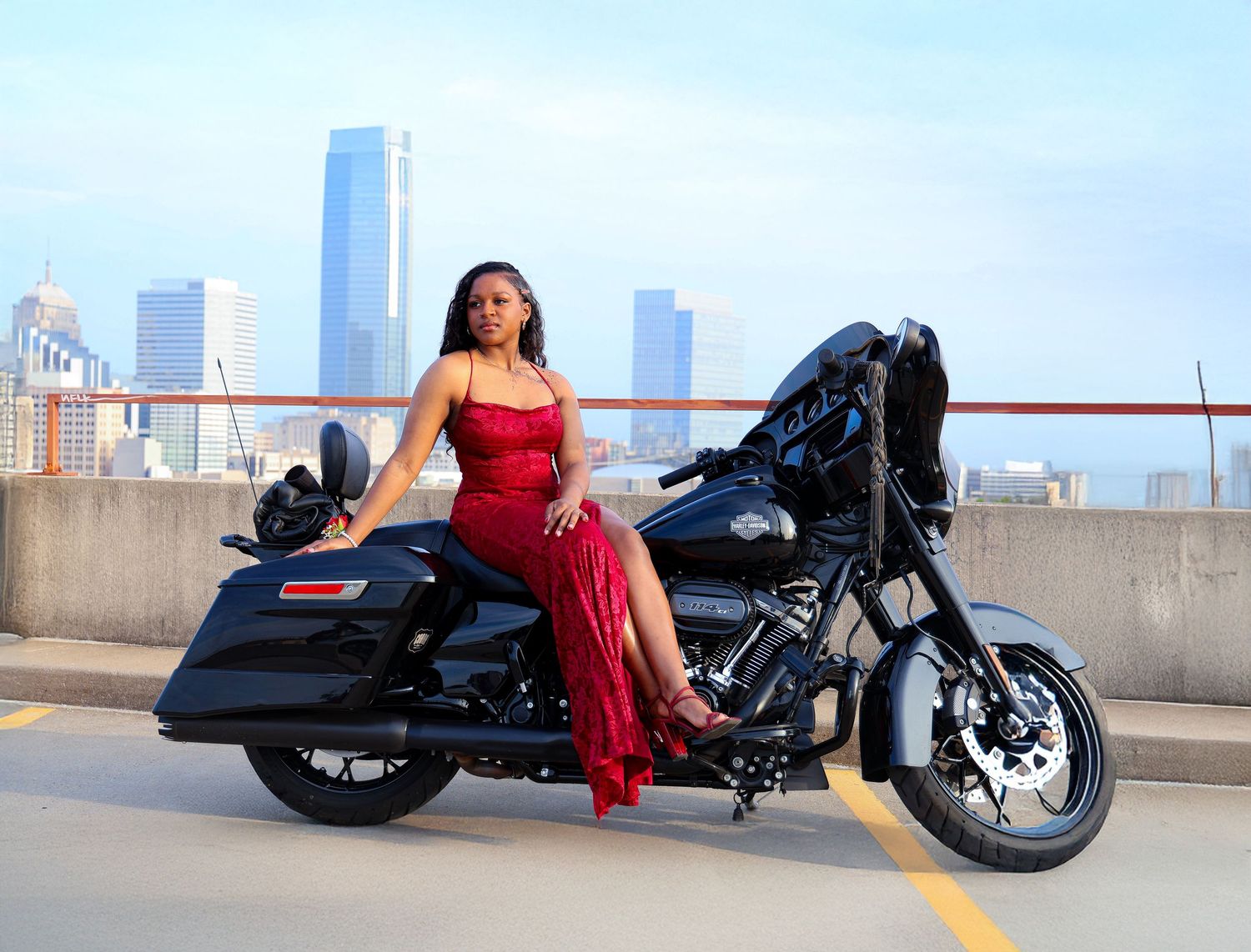 Woman in red dress poses on black Harley-Davidson motorcycle on rooftop with city skyline backdrop.
