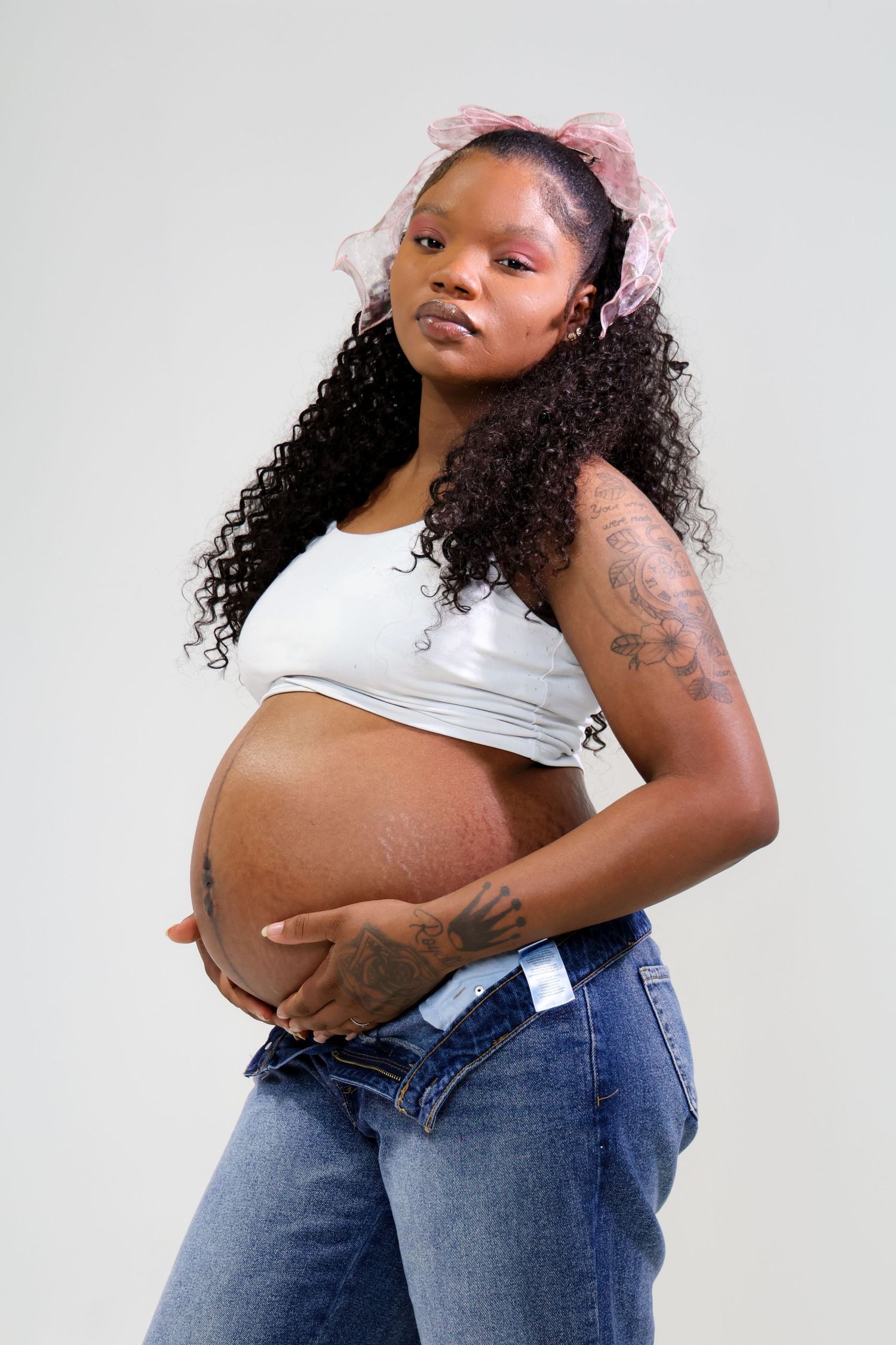 Pregnant Black woman with curly hair and pink bow cradling baby bump, wearing white crop top and jeans.