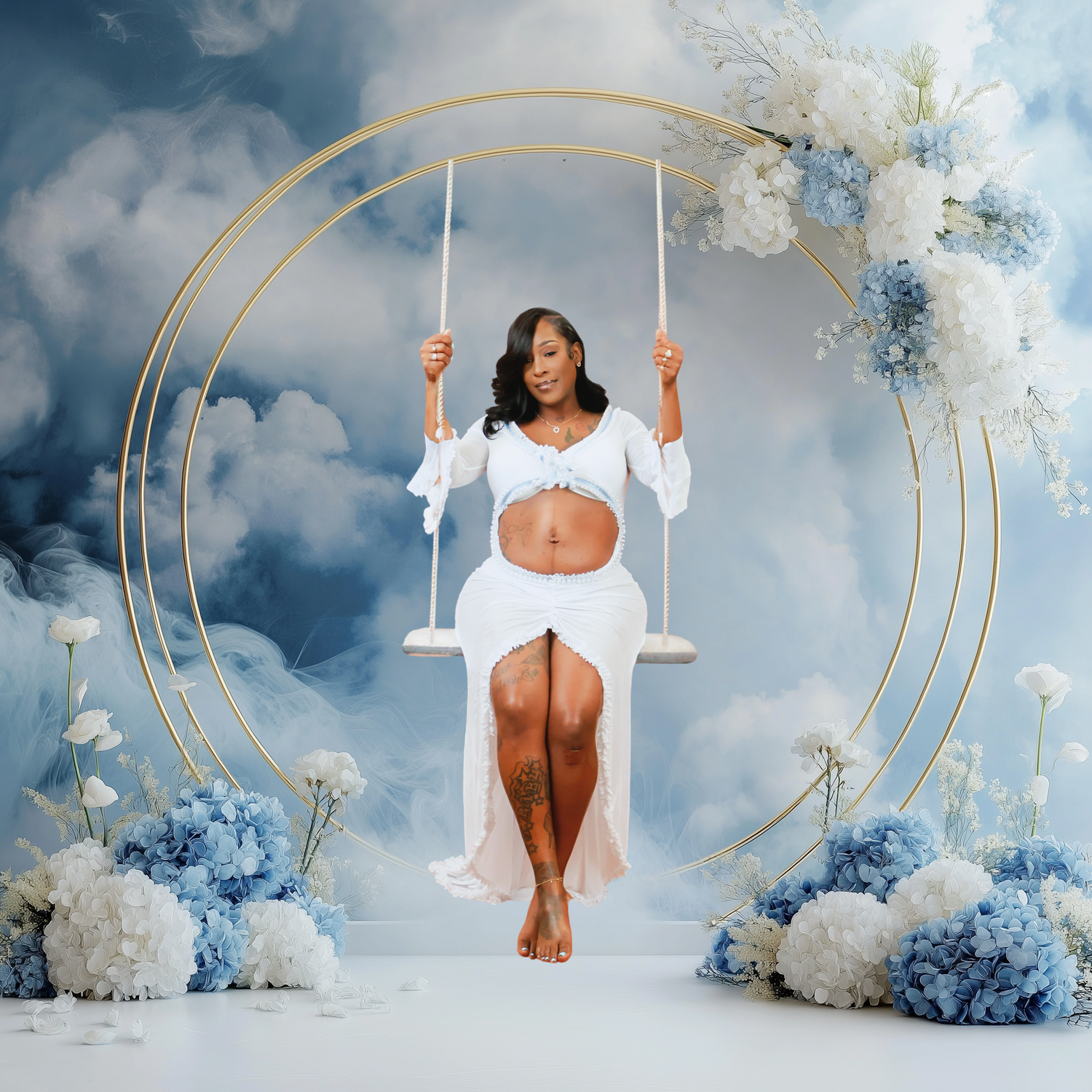 Pregnant woman in white outfit sitting on gold swing surrounded by blue and white floral arrangement on cloudy backdrop.
