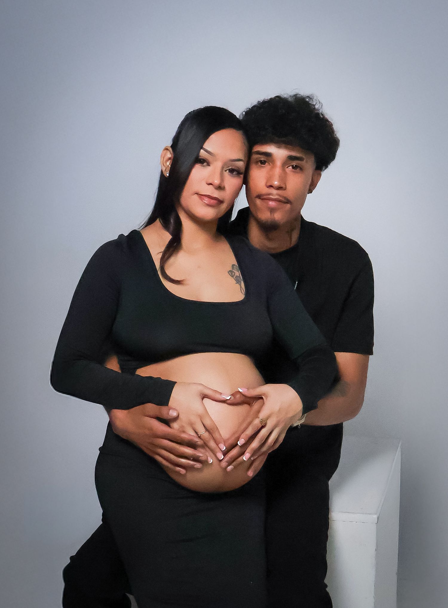 Couple in black outfits posing for maternity photo, man embracing pregnant woman forming heart shape on belly.